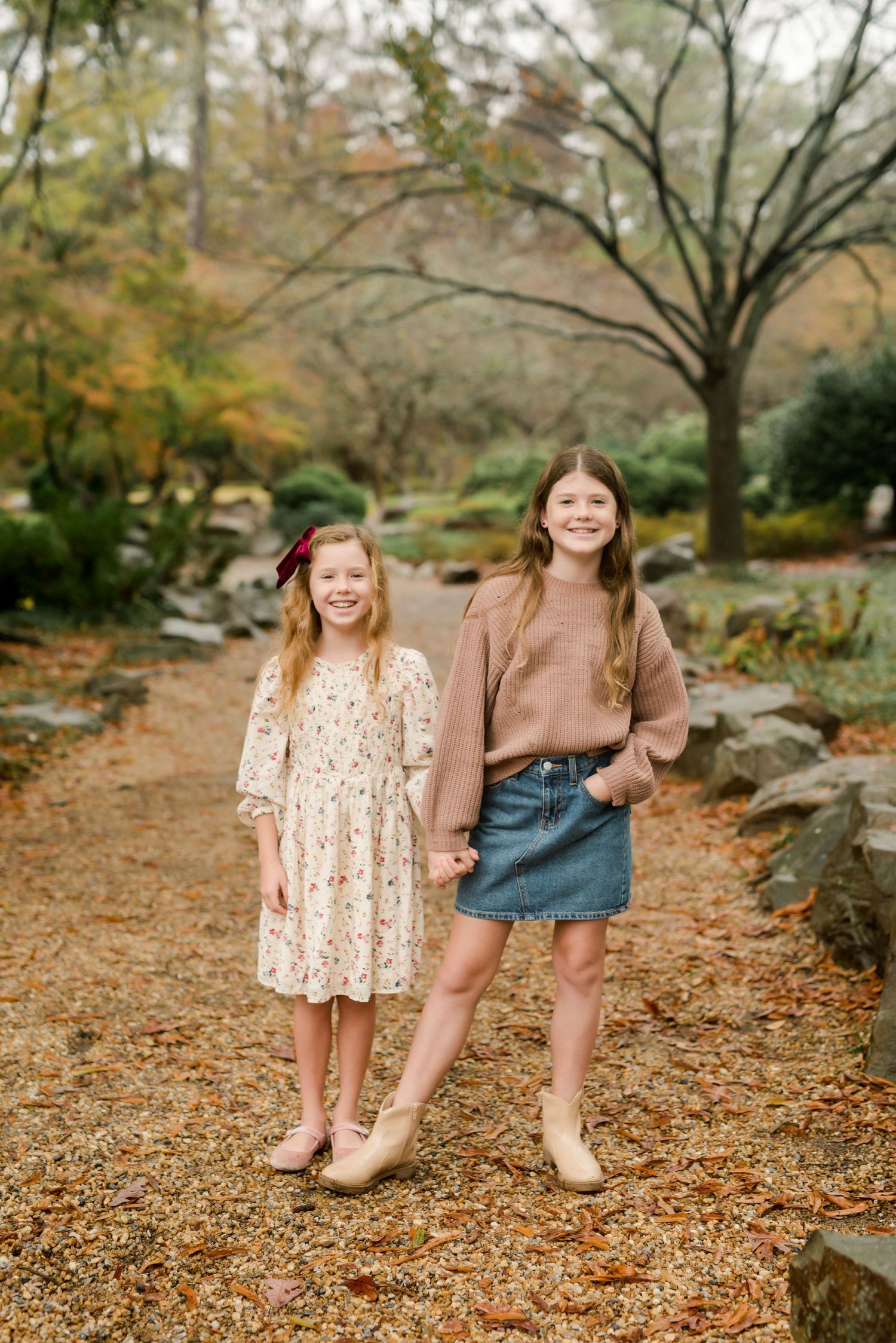 Two people hold hands while standing on leaf-covered path in autumn garden setting.