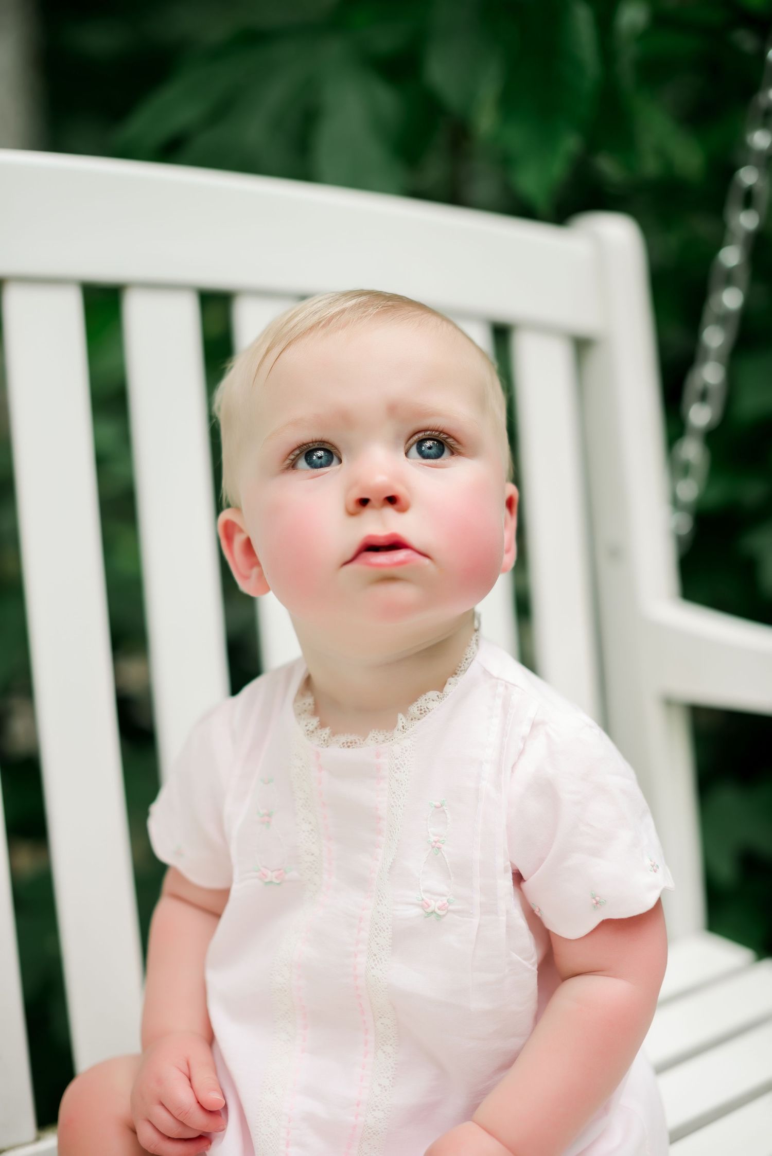 Close up portrait of baby in white outfit sitting on white garden chair.