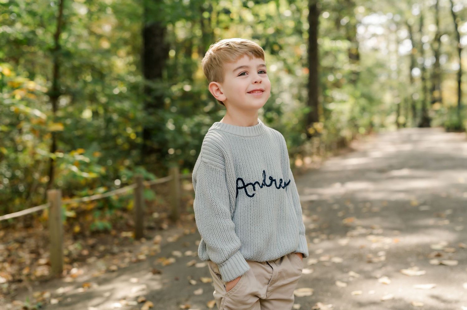 Young boy wearing a gray personalized knit sweater with name Andrew, standing on a forest path.