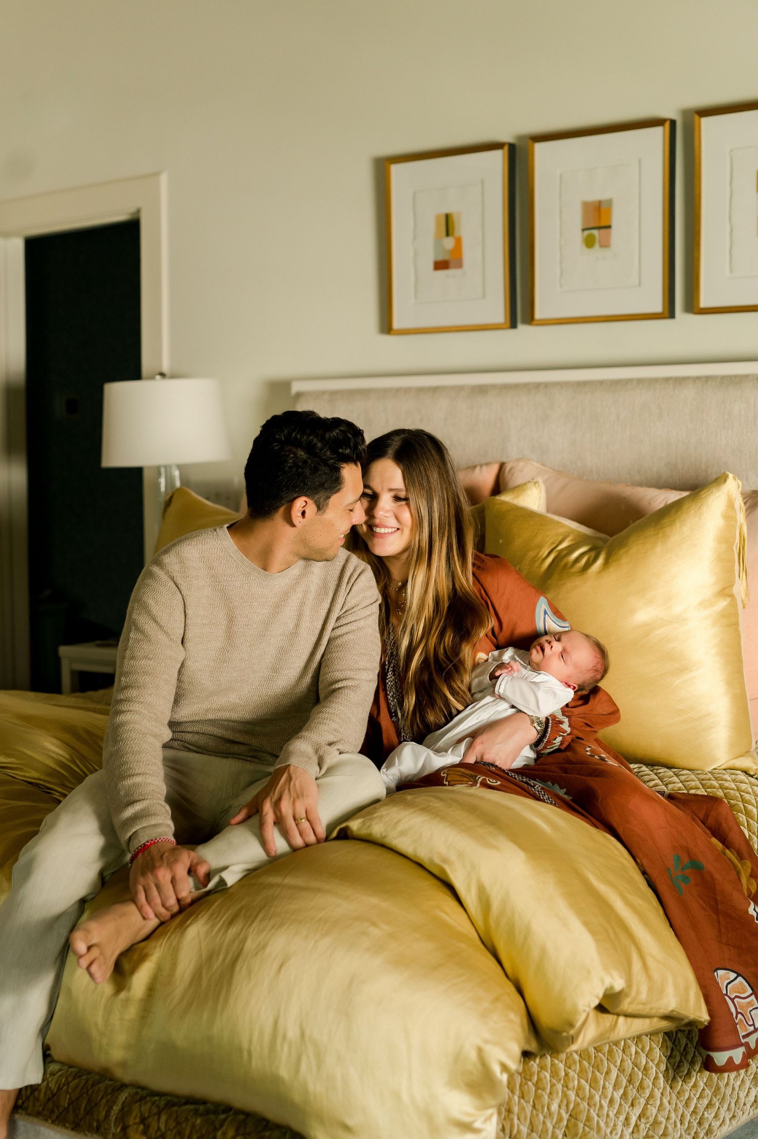 A couple sits on a golden bed with their newborn baby, sharing a tender moment in a warmly decorated bedroom.