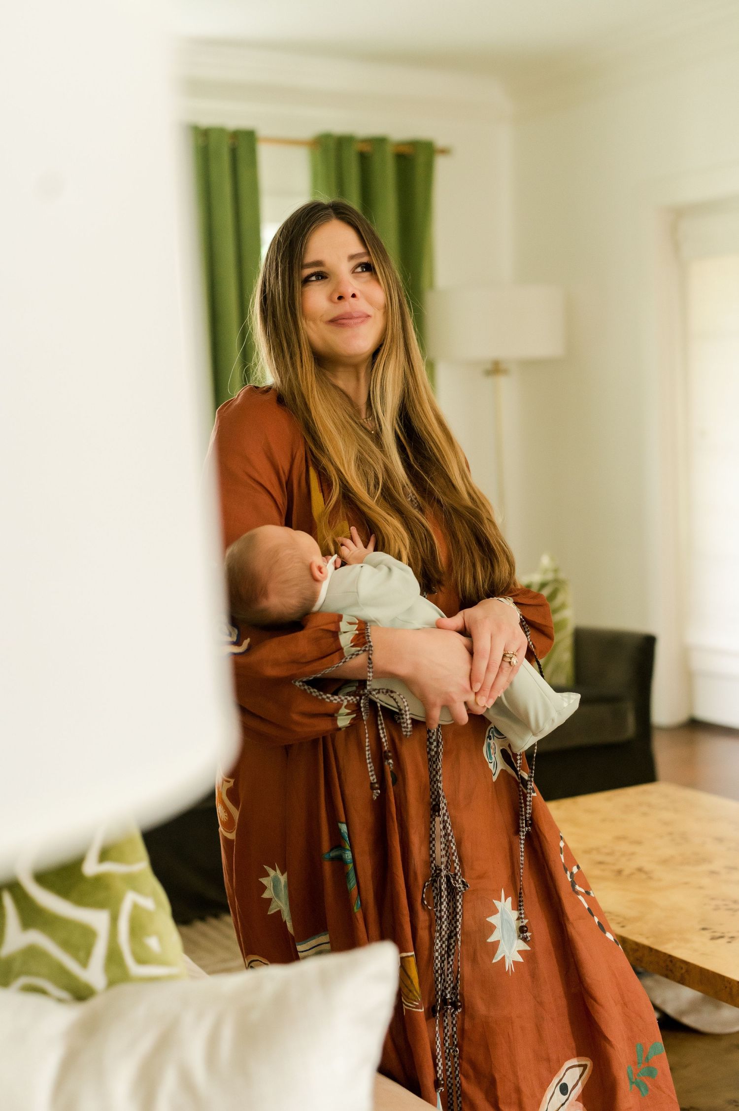 A young mother in a rust boho dress holds her newborn baby while standing in a bright living room.