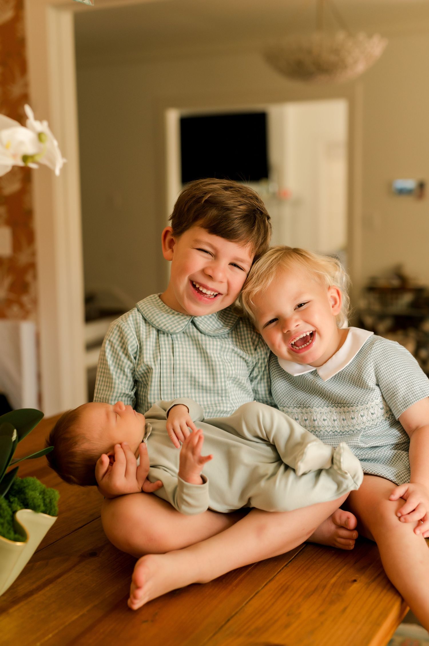 Two smiling boys sitting on a table holding a sleeping newborn baby sibling indoors.