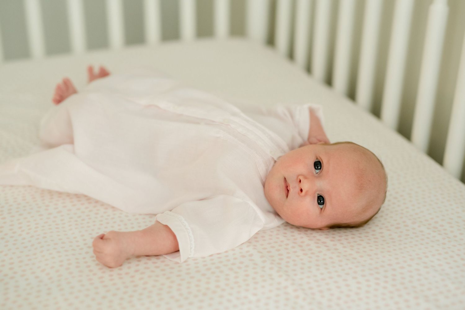 Newborn baby in white onesie lying on white crib mattress.