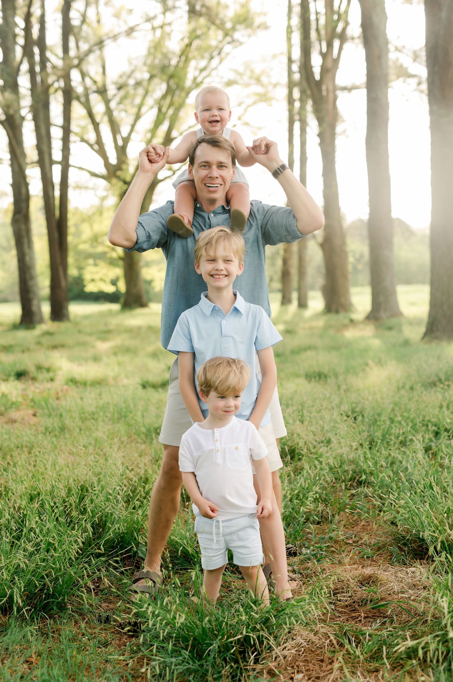 A parent carries a toddler on shoulders while standing outdoors with two other children in a sunlit wooded area.