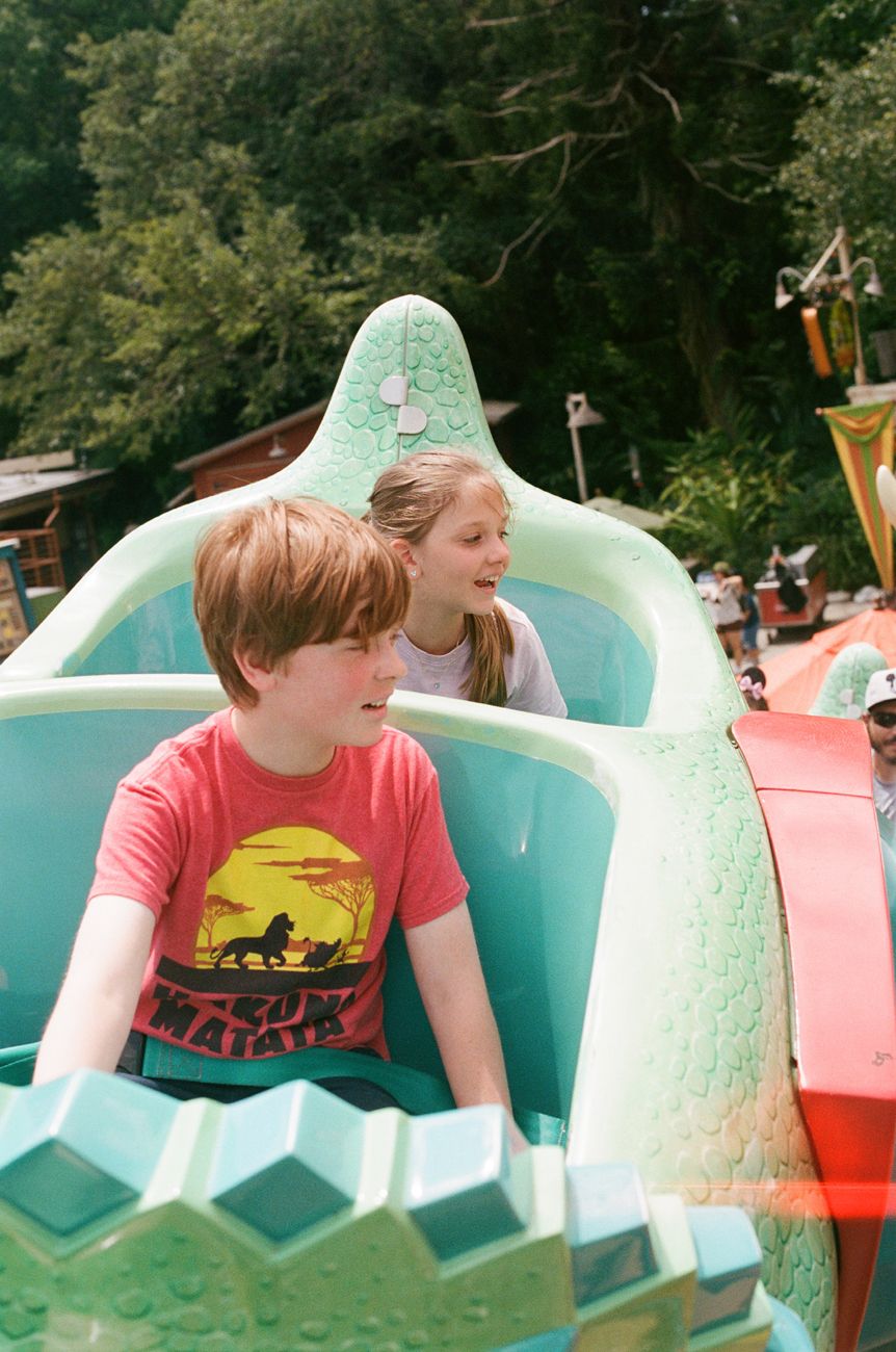 Young riders enjoy a colorful dinosaur-themed amusement park ride on a sunny day.