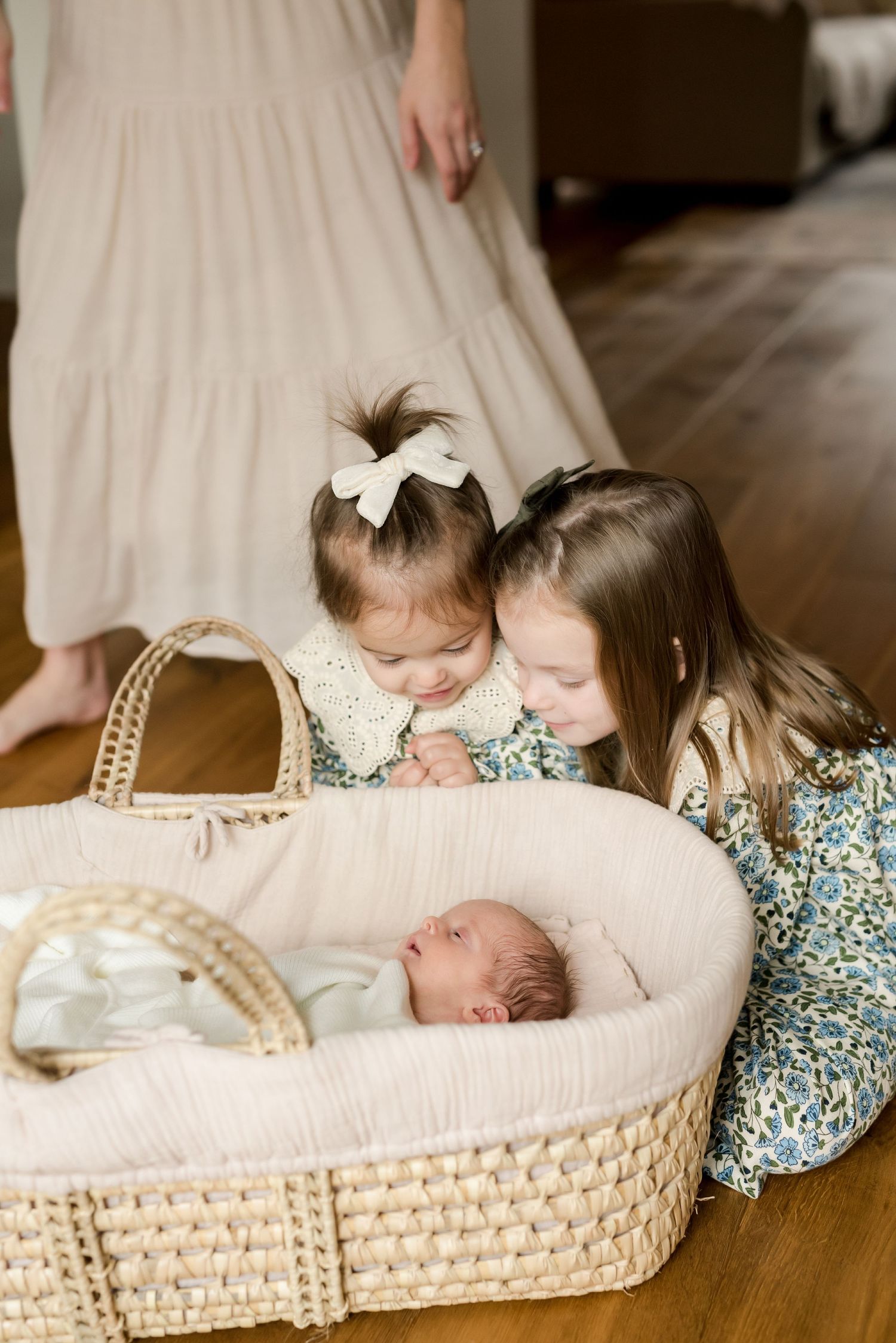 Children peering into woven bassinet at newborn baby on hardwood floor.