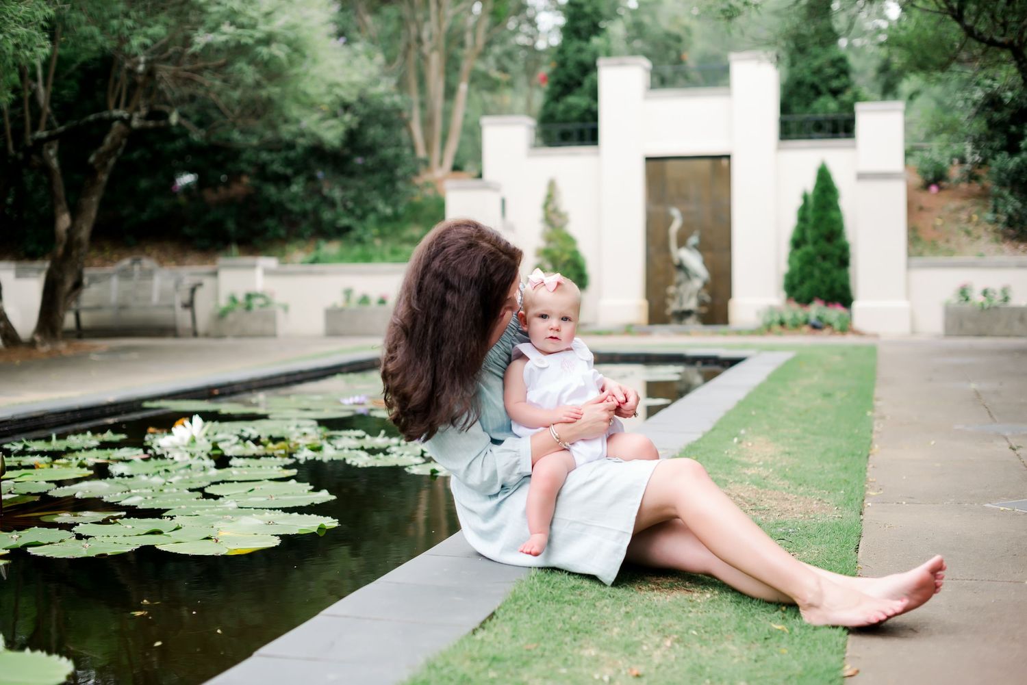 Mother and baby sit by a serene lily pond in a formal garden with white architectural features.