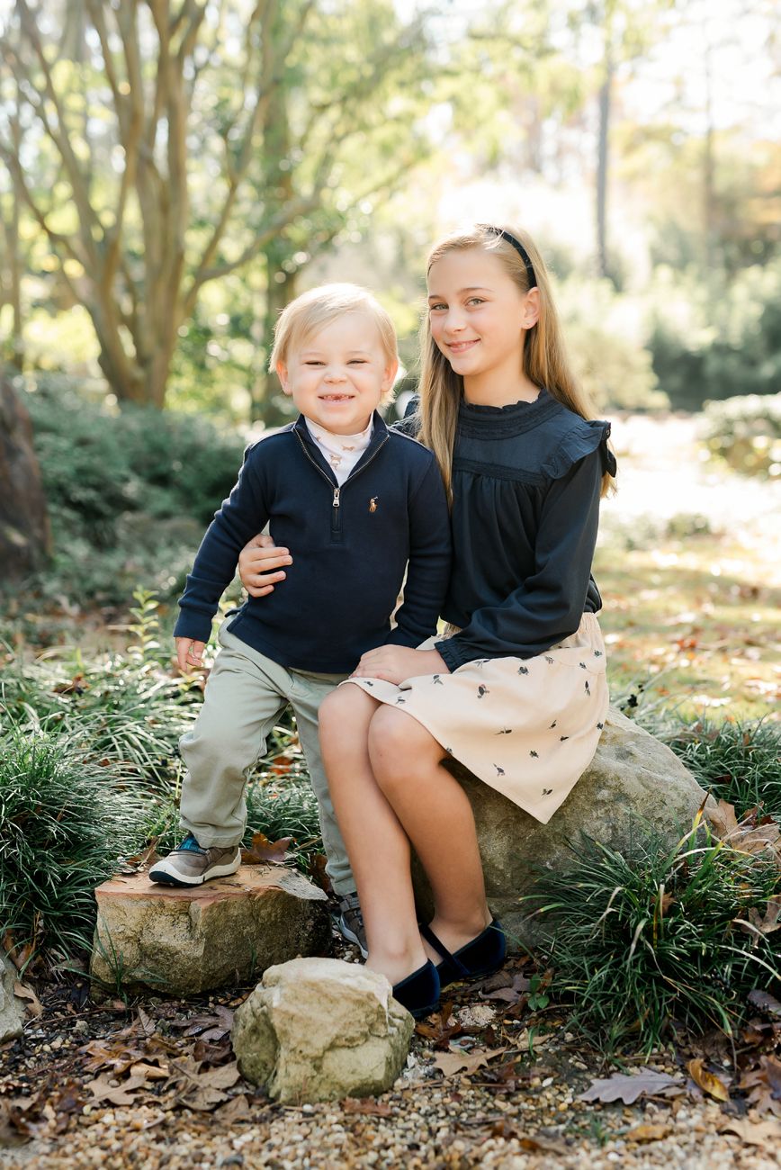 Two children pose on rocks outdoors, a smiling boy in navy and a girl in black top with tan skirt, in a wooded setting.