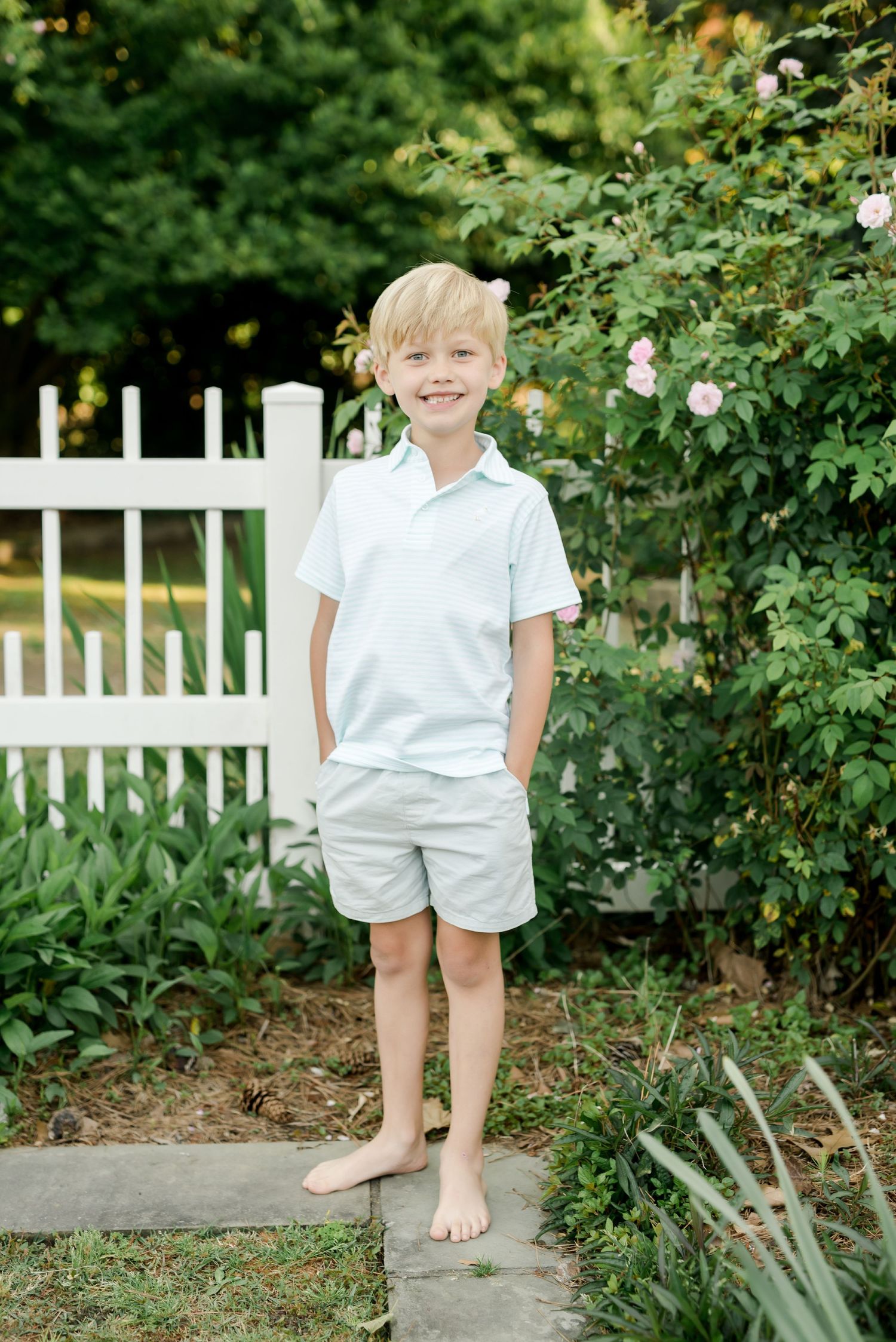 Young boy in white polo and khaki shorts poses by white picket fence and garden.