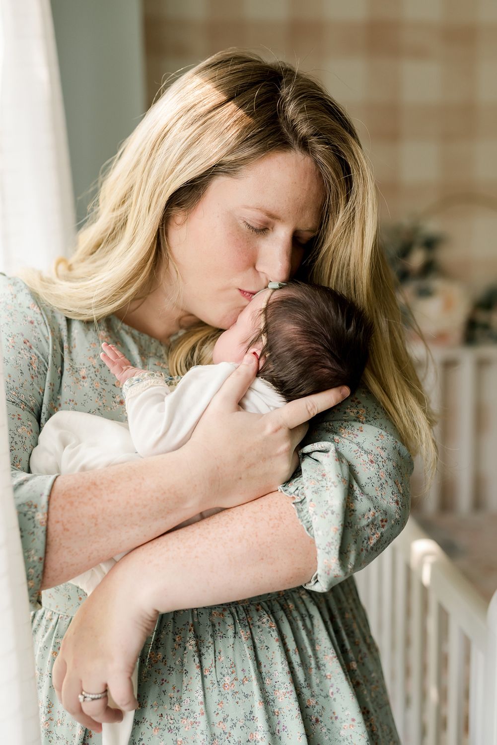 Woman in floral dress holding and tenderly kissing a small infant in a bright interior setting.