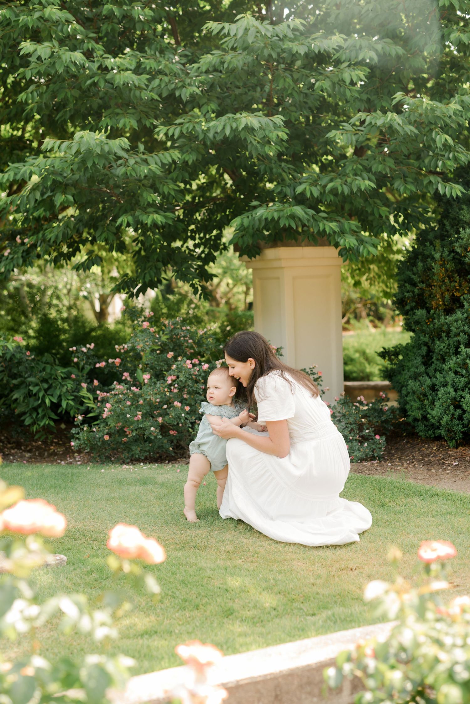 Mother and baby sharing tender moment in garden with pink flowers and white column.