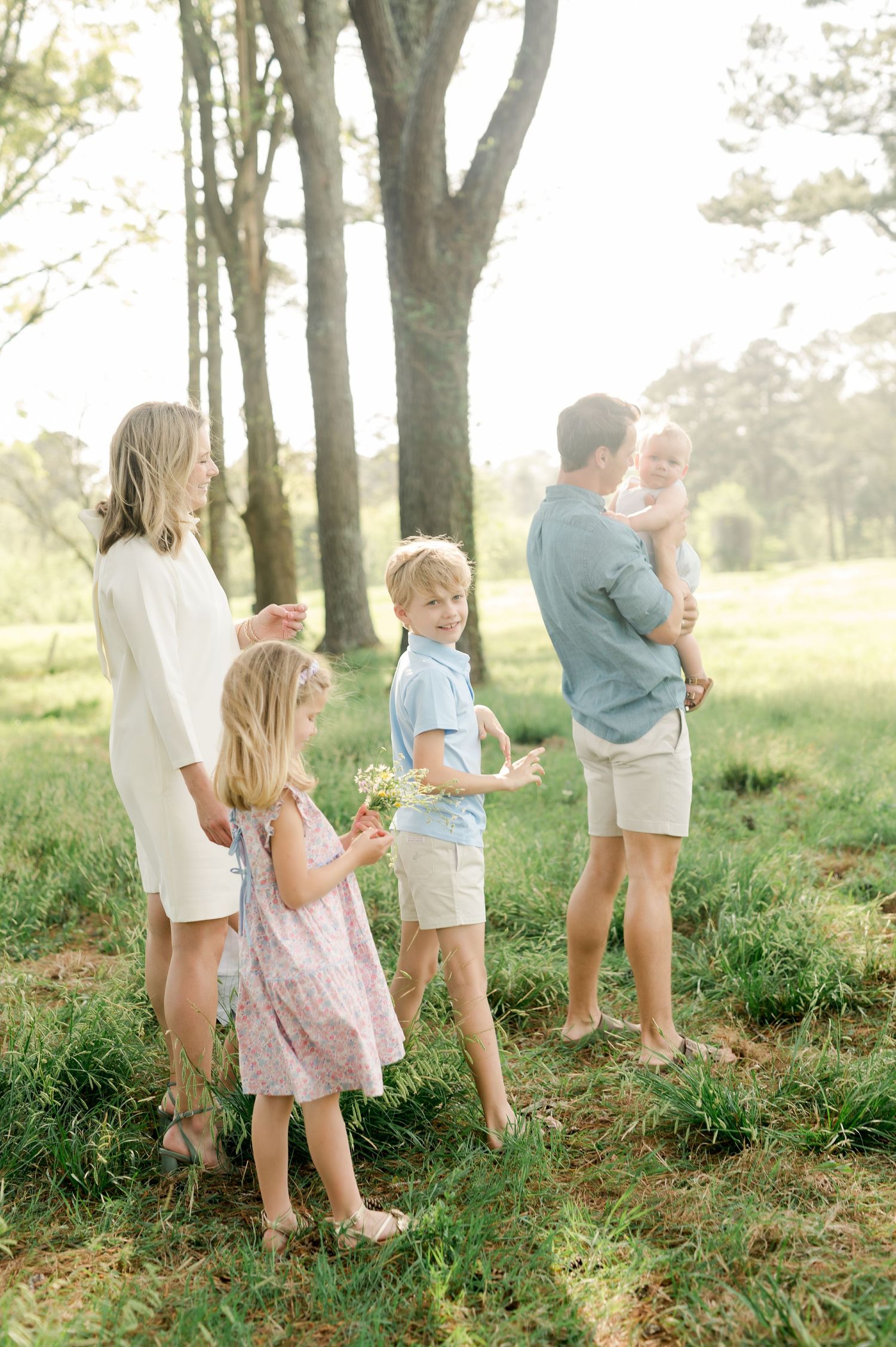 A family stands together outdoors in a sun-dappled wooded area wearing light summer clothing.