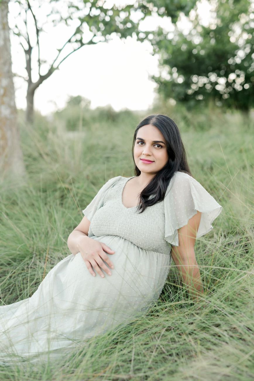 Expectant mother in a flowing white dress poses gracefully in tall grass during outdoor maternity session.