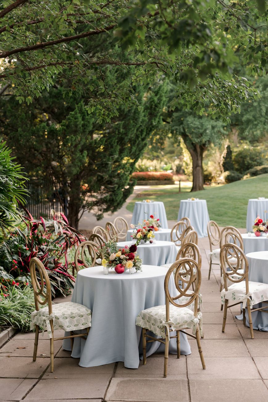Elegant outdoor event setup with round tables covered in white linens and gold chairs under trees.