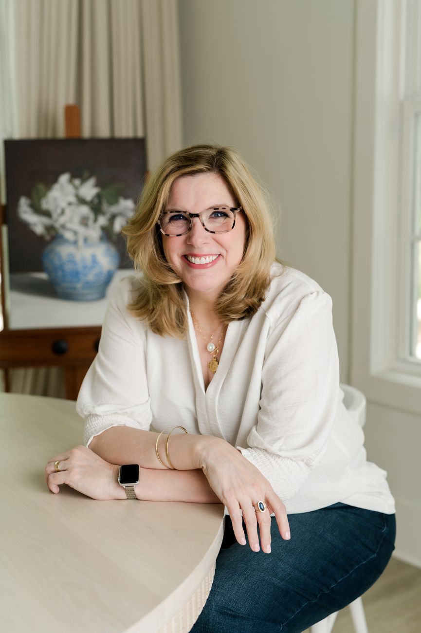 Person in white blazer and glasses poses casually at desk.