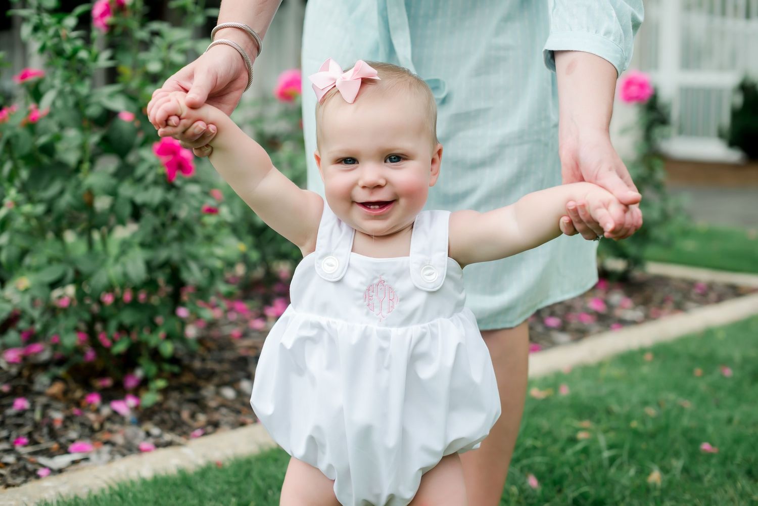 Happy baby in white outfit smiles while being supported standing in a blooming garden.