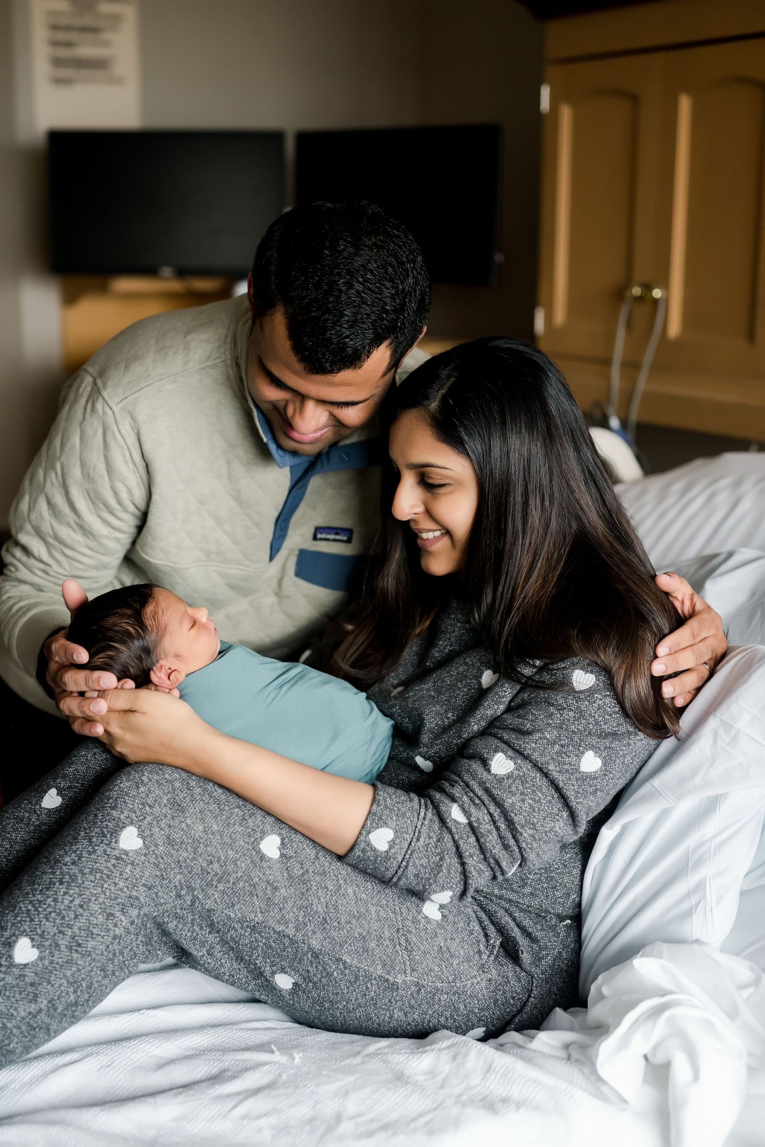 Parents share a loving moment with their newborn baby while resting on a hospital bed.