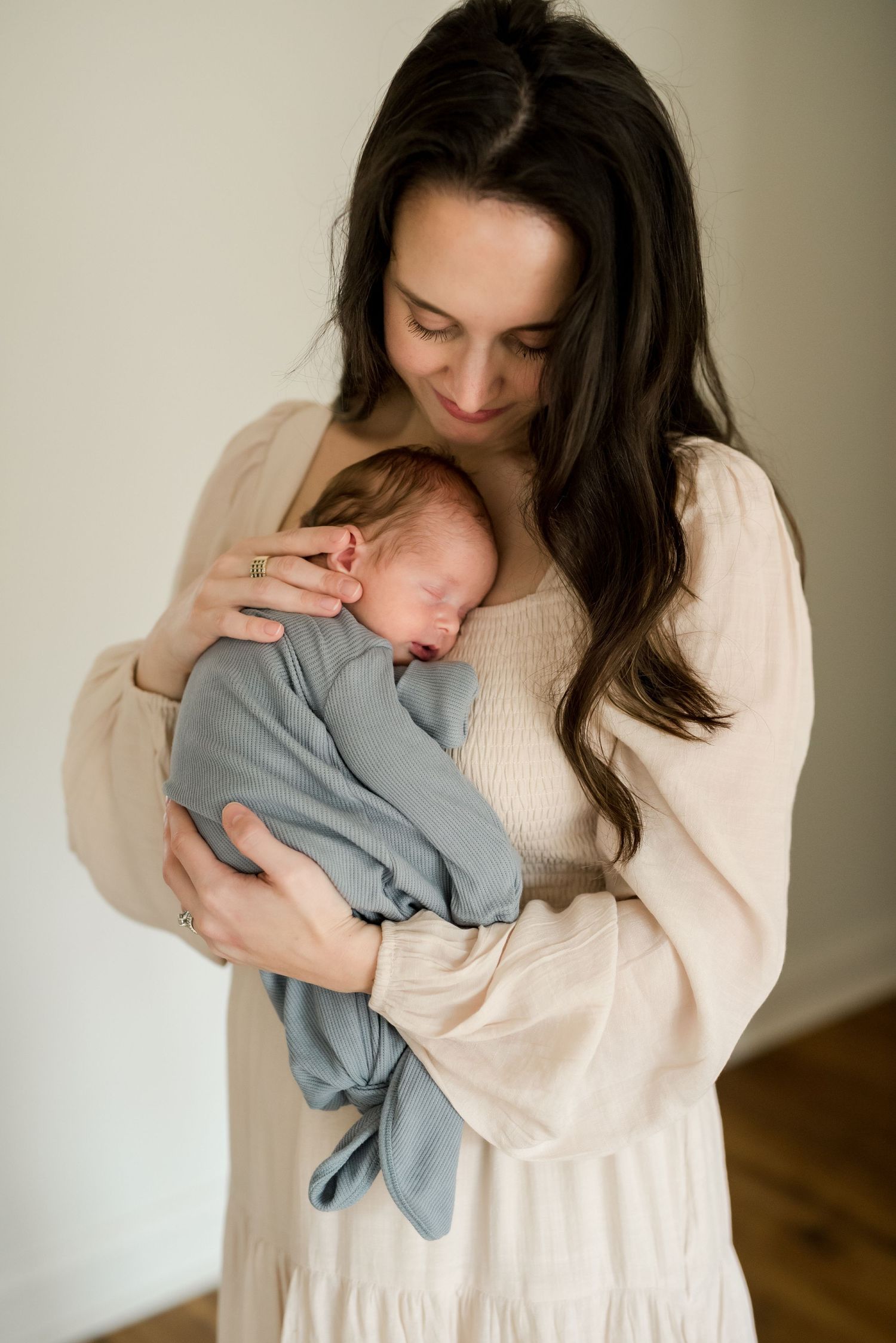 Mother in cream sweater holding sleeping newborn baby dressed in soft grey clothing.