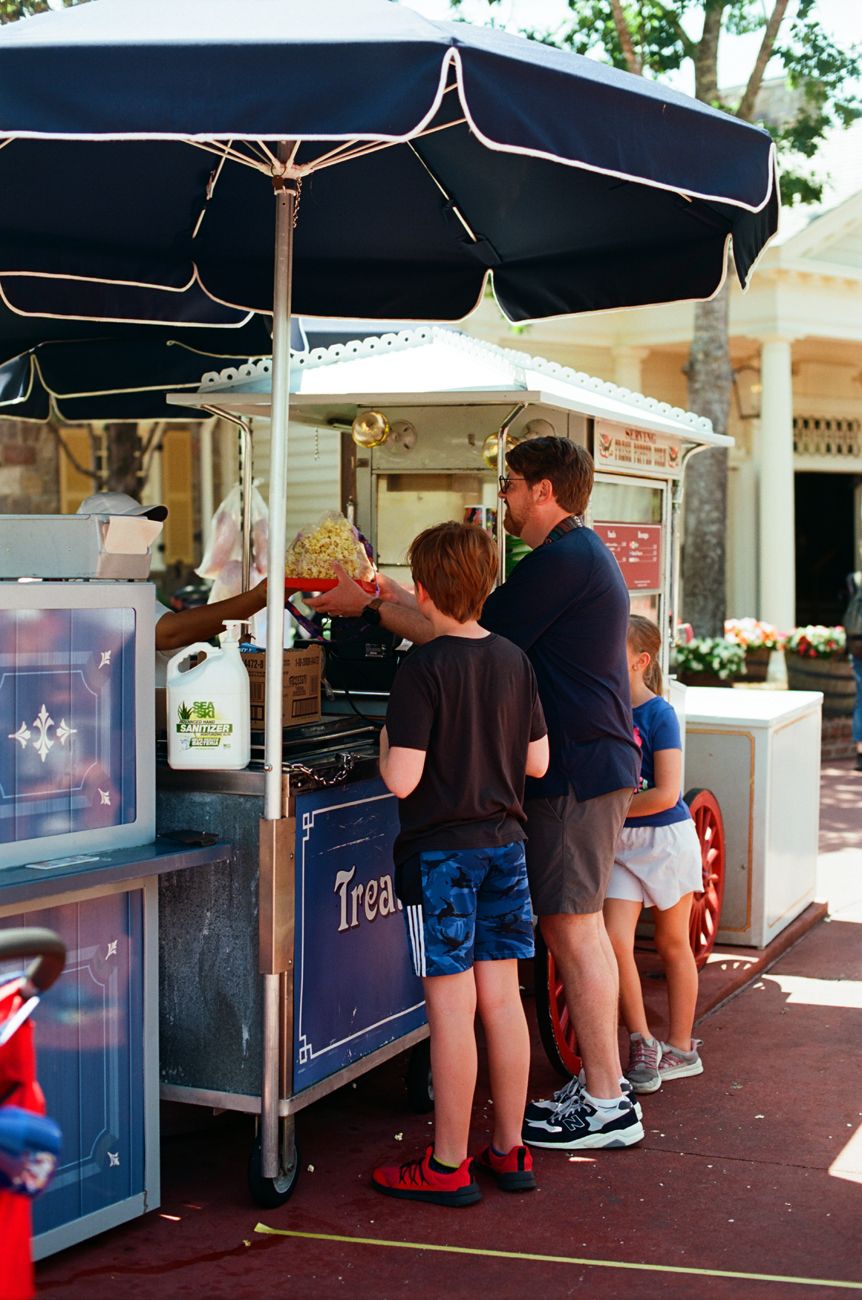 Visitors ordering from a snack kiosk under black umbrellas at a theme park.