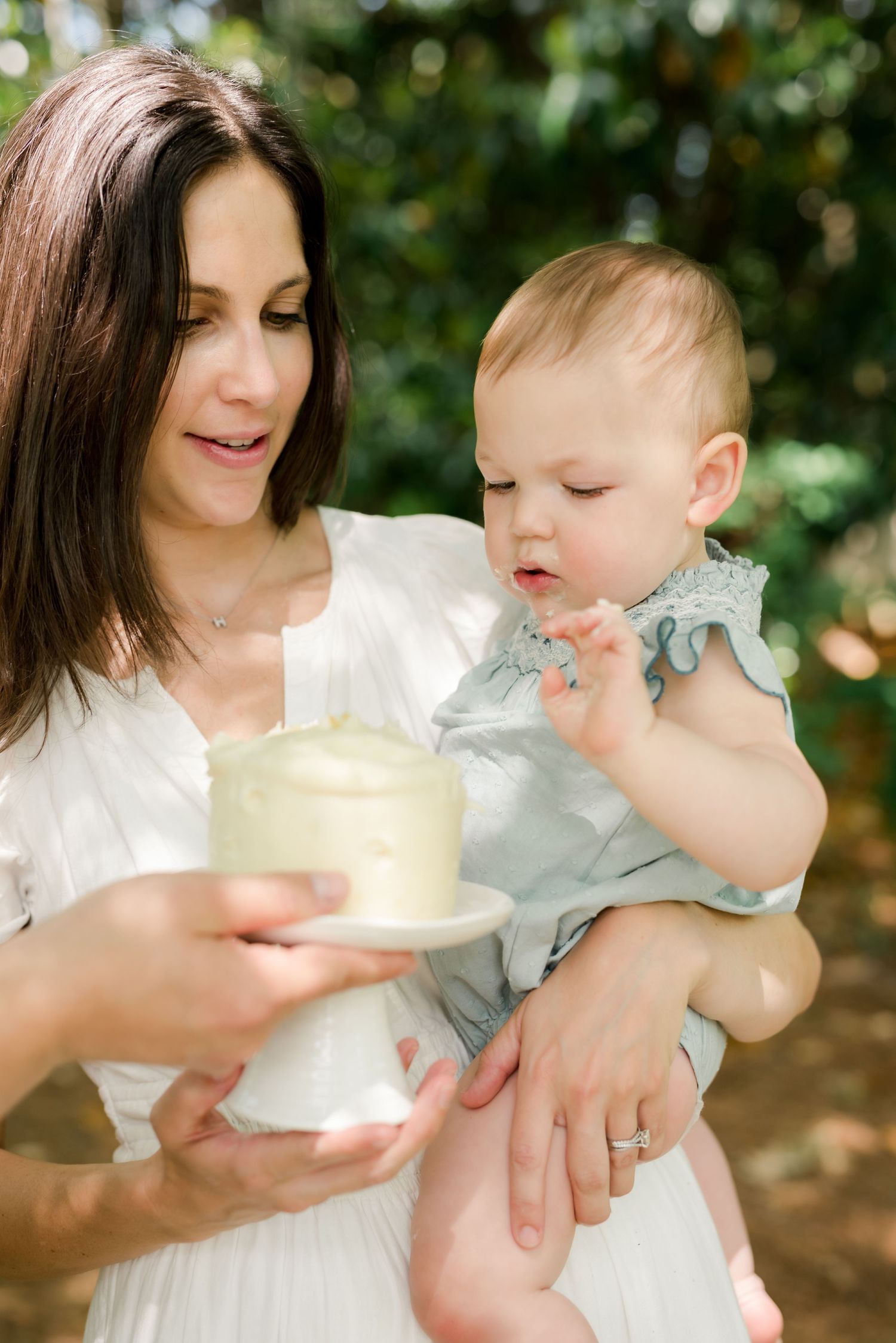 Mother holding baby while offering cake in garden setting.