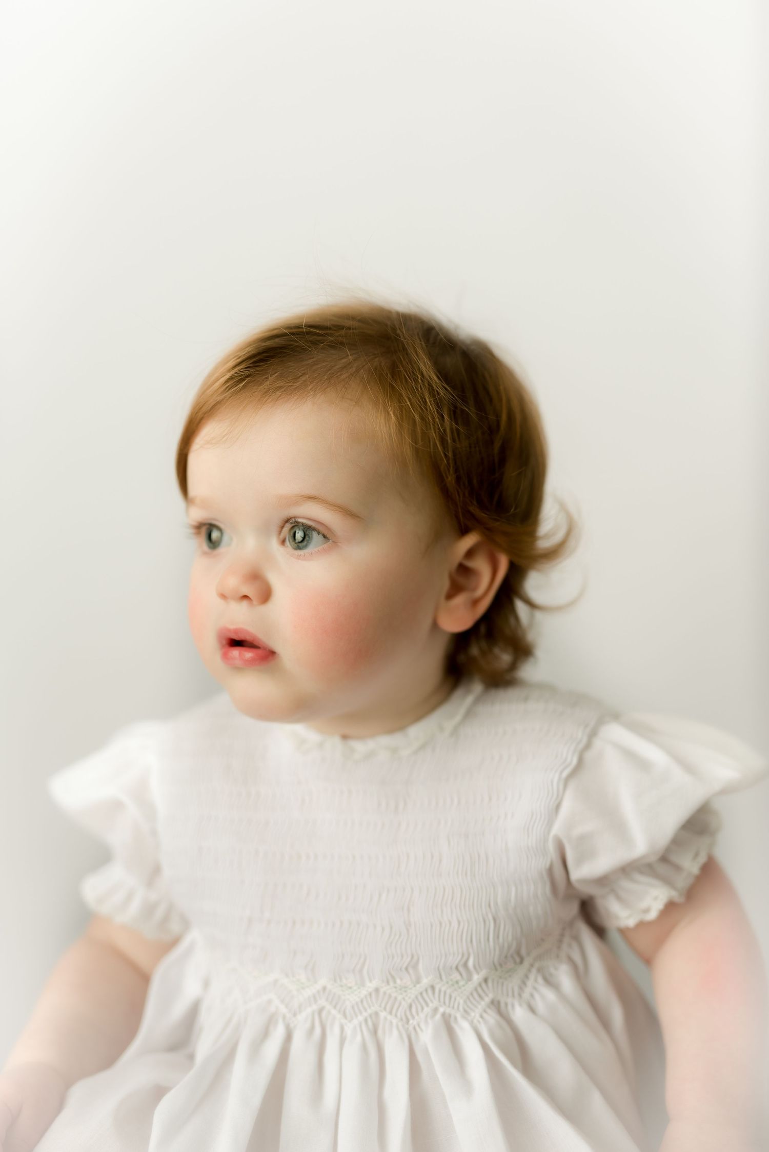 Baby wearing white smocked dress looking to side against white backdrop.
