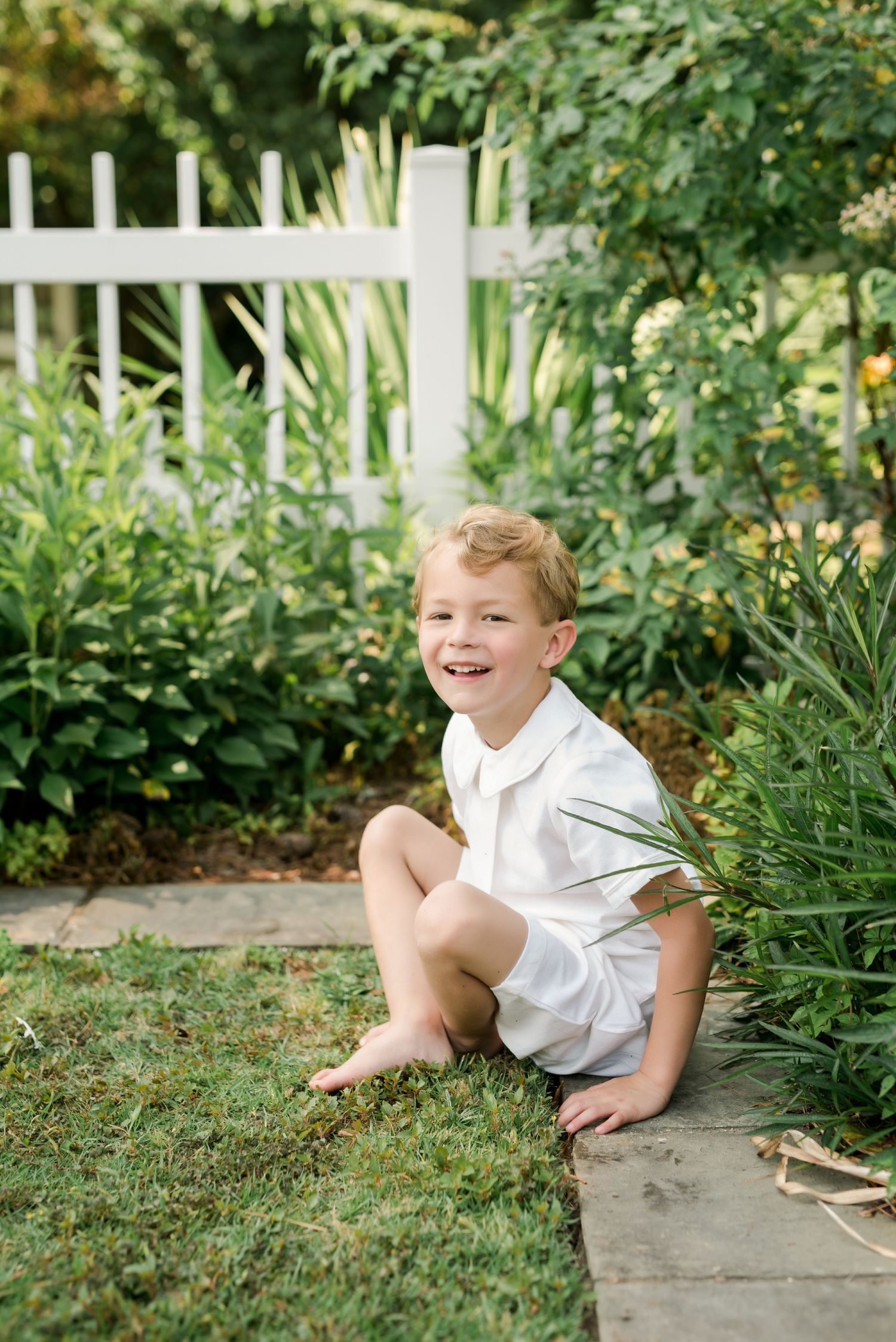 Child in white outfit sits playfully on garden path edge surrounded by greenery.