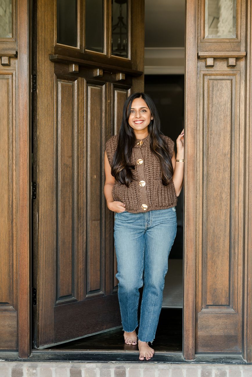 Person in brown sweater and jeans posing against wooden paneled doorway.