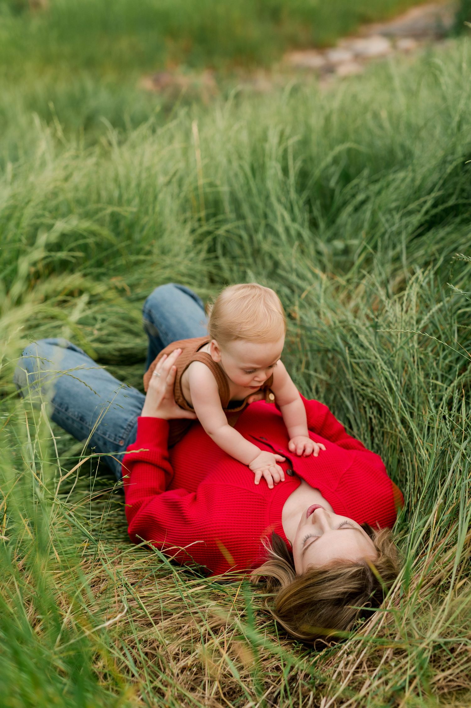 A mother in a red coat lies in tall green grass, smiling as her baby crawls on her chest outdoors.