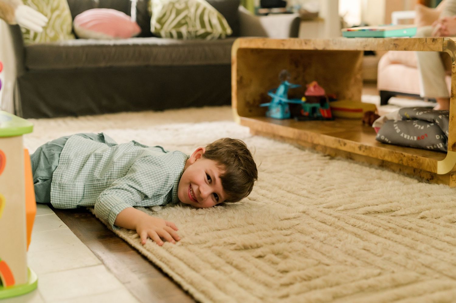 Young boy lying on a beige carpet in a cozy living room, smiling while playing on the floor.