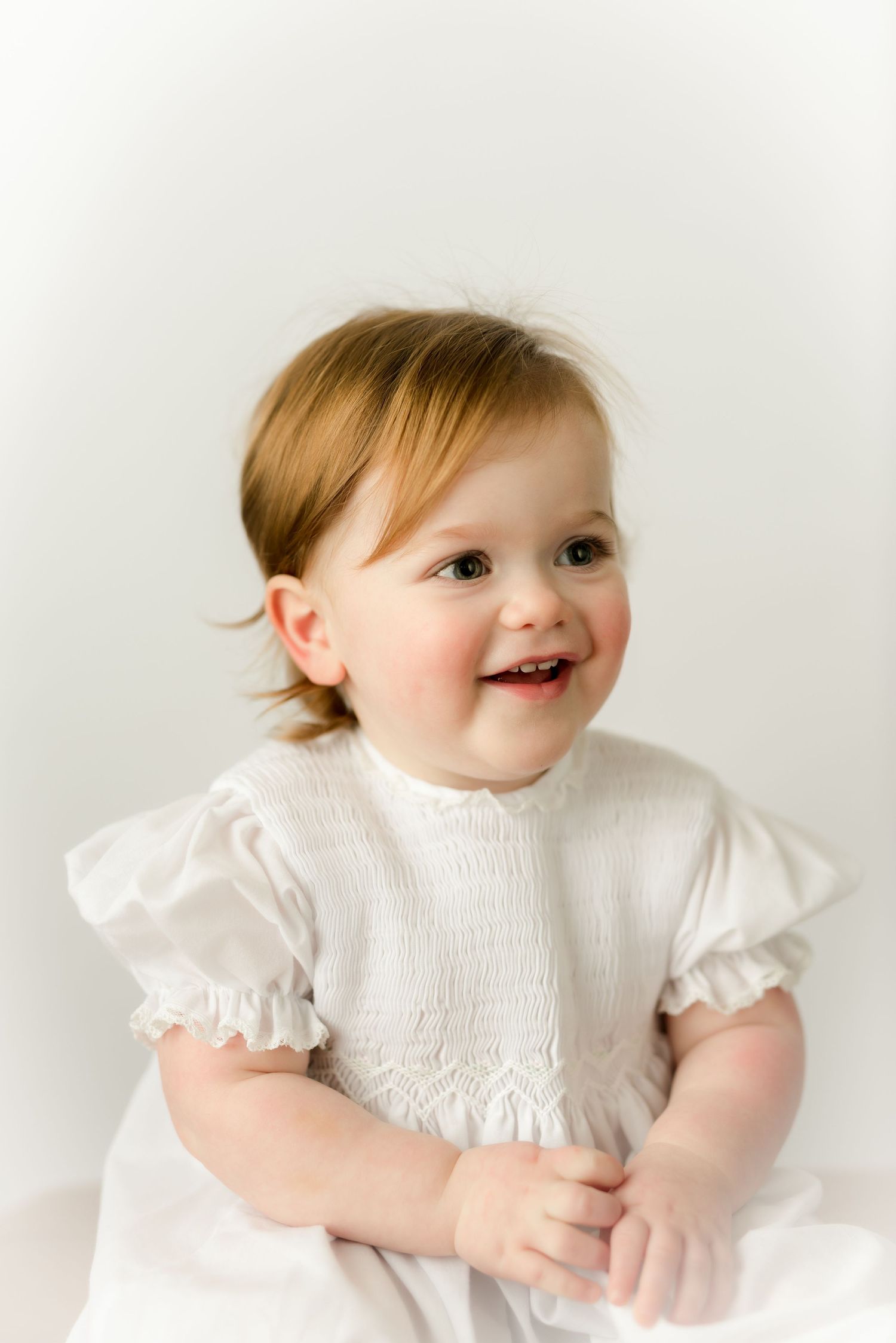 Baby in white smocked dress smiling against white background.