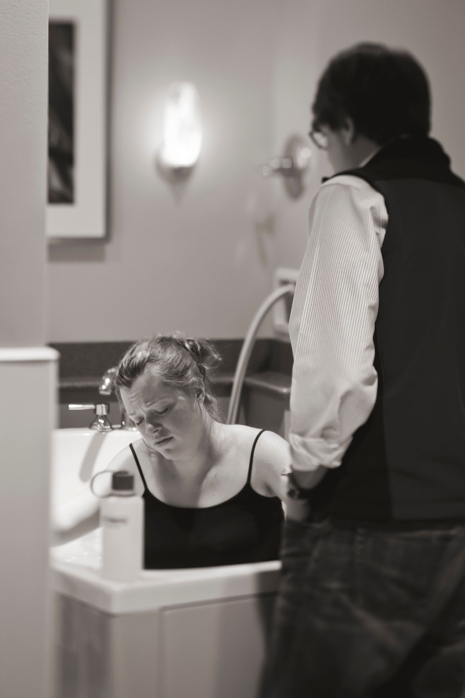 Black and white image showing person bent over bathroom sink with another figure standing nearby.