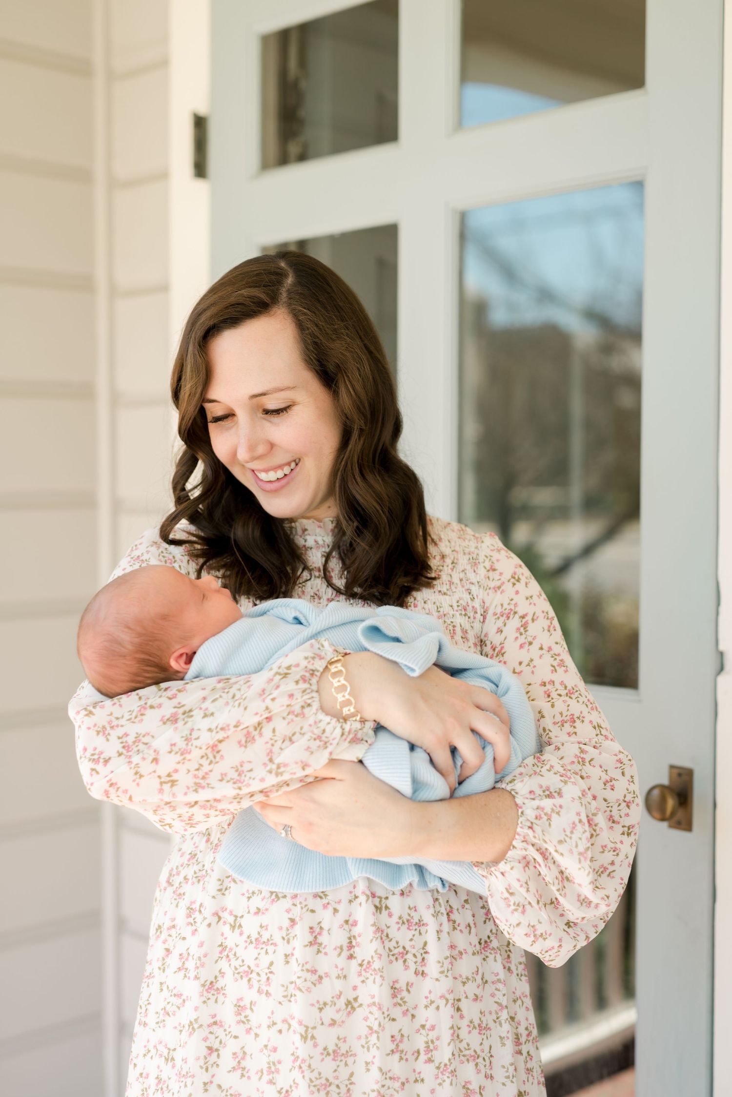 Mother in floral dress holding sleeping newborn baby on front porch.