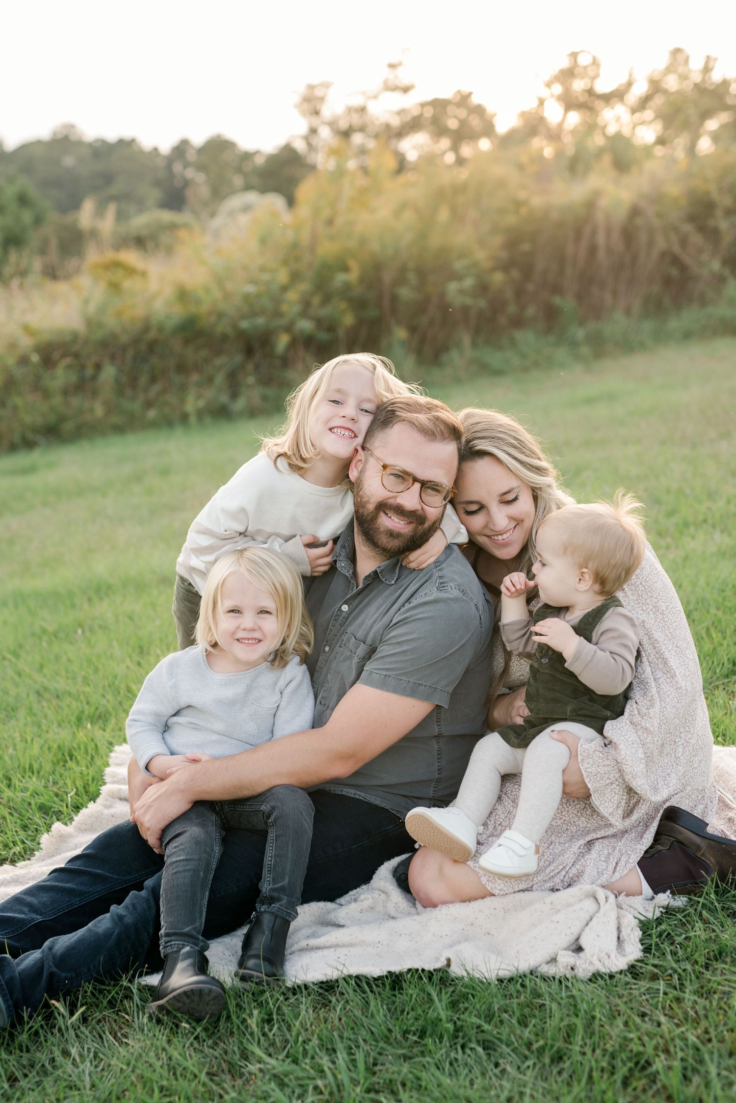 Family cuddle moment captured on blanket in grassy field during sunset.