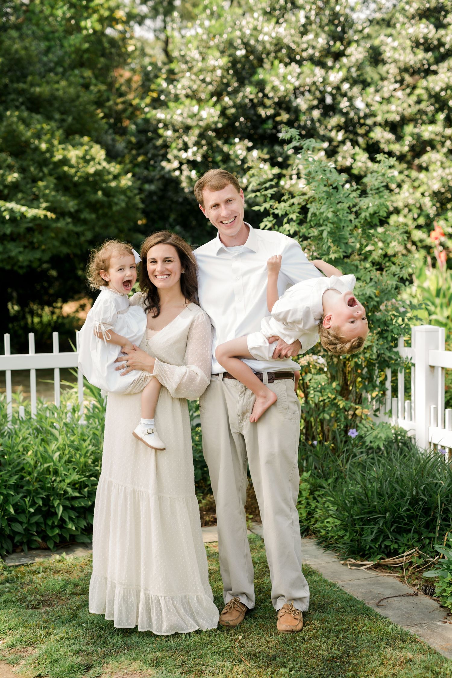 A family dressed in white and cream tones poses together in a garden setting with a white fence.