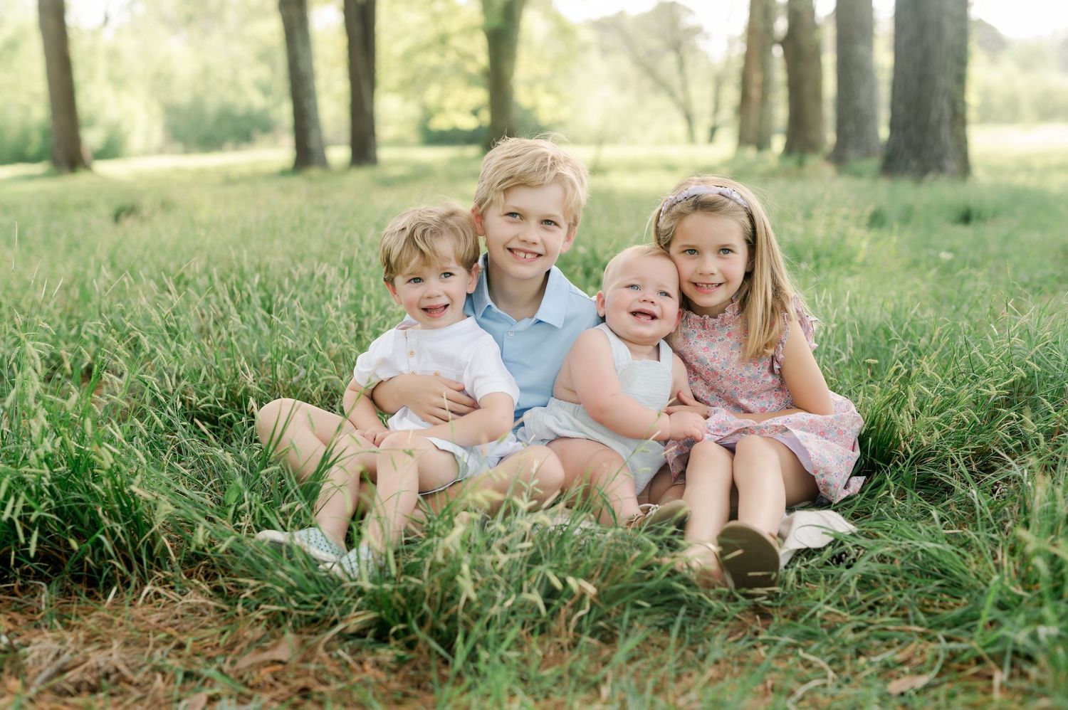 Four young children sitting together in tall grass under a row of trees on a sunny day.