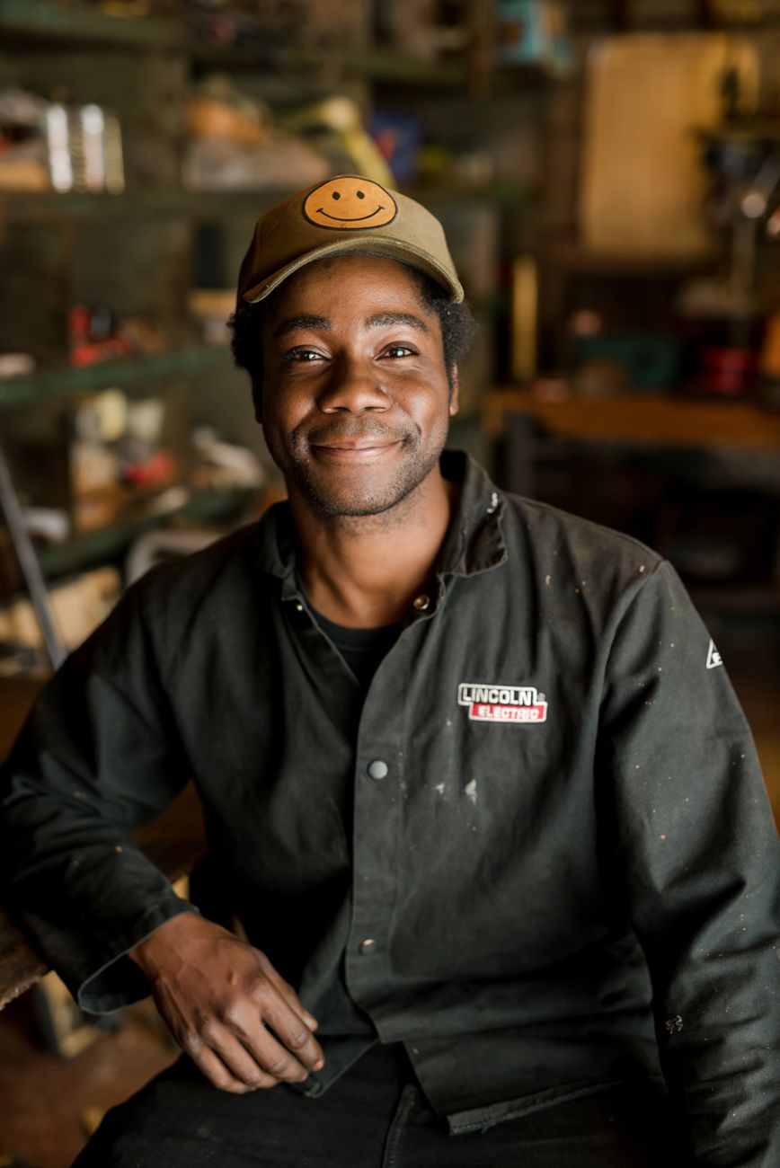 Person in dark workwear and cap smiling warmly at camera in dim lighting.