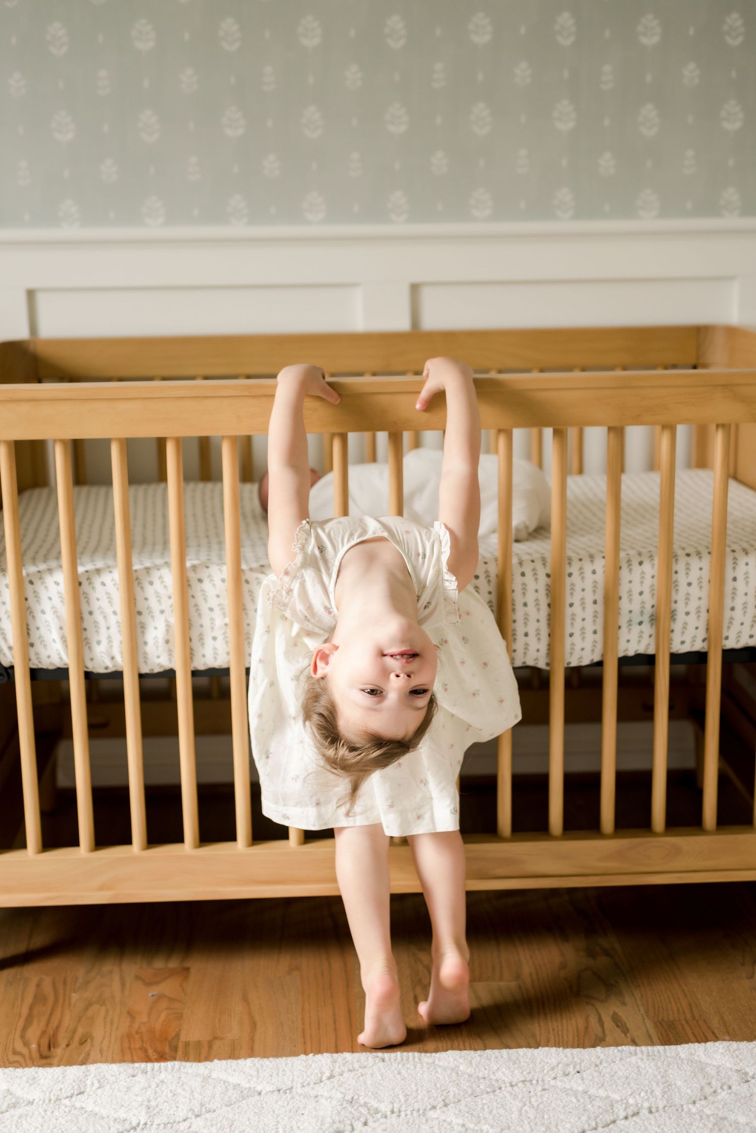 A playful child in a white dress hangs upside down from a wooden crib while smiling.