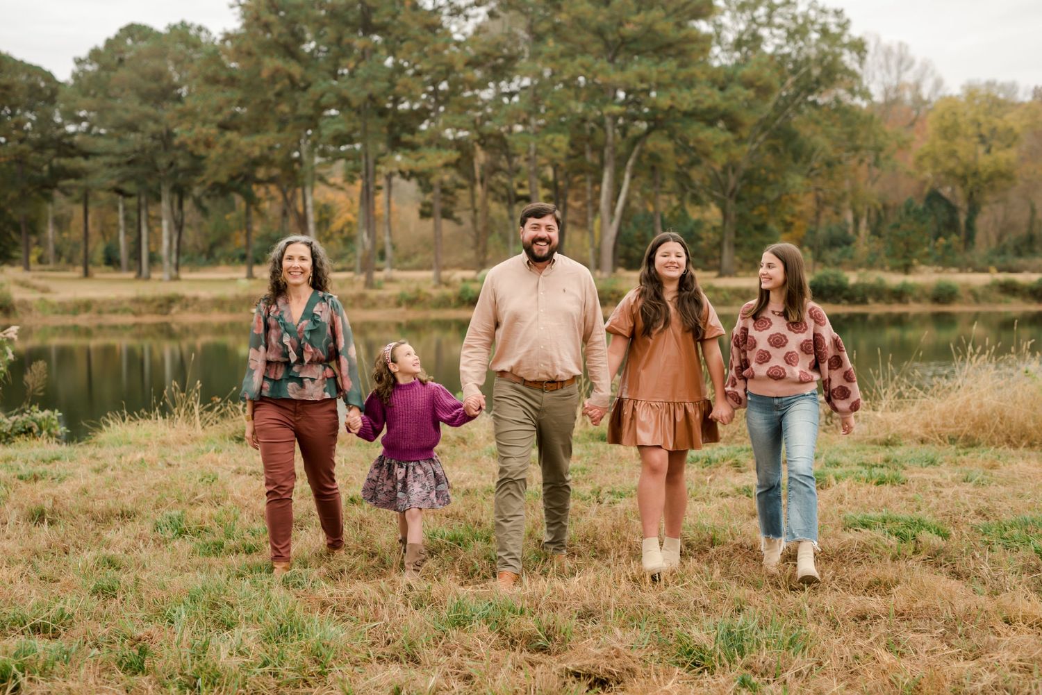 Family walking together through autumn field near lake with pine trees in background.
