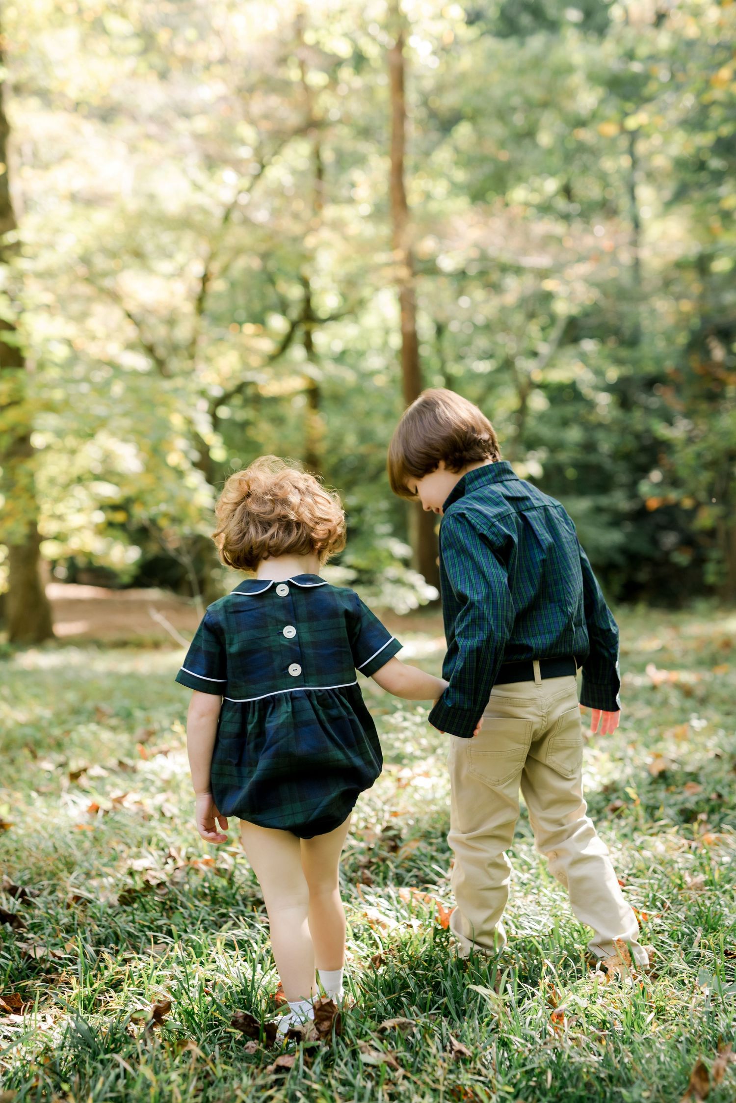 Two children walk together through a sunlit forest wearing coordinated navy outfits on a fall day.