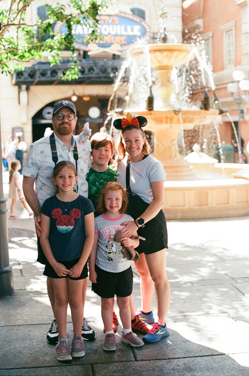 A group posing together in front of a fountain at a theme park wearing Disney-themed clothing and accessories.