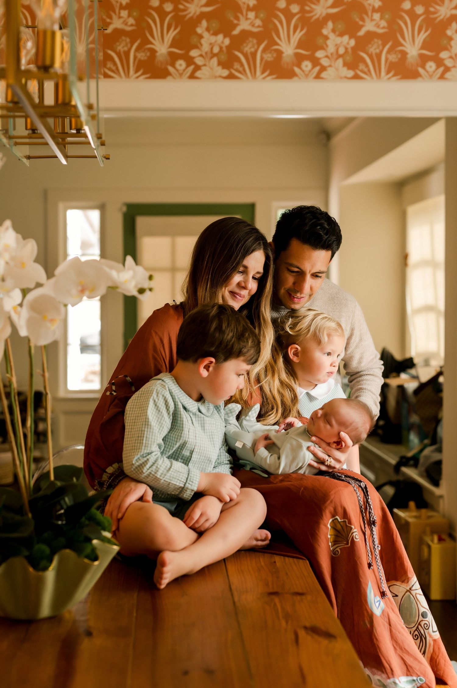 A warm family portrait with parents and three young children, including a newborn, gathered together in a cozy home interior.