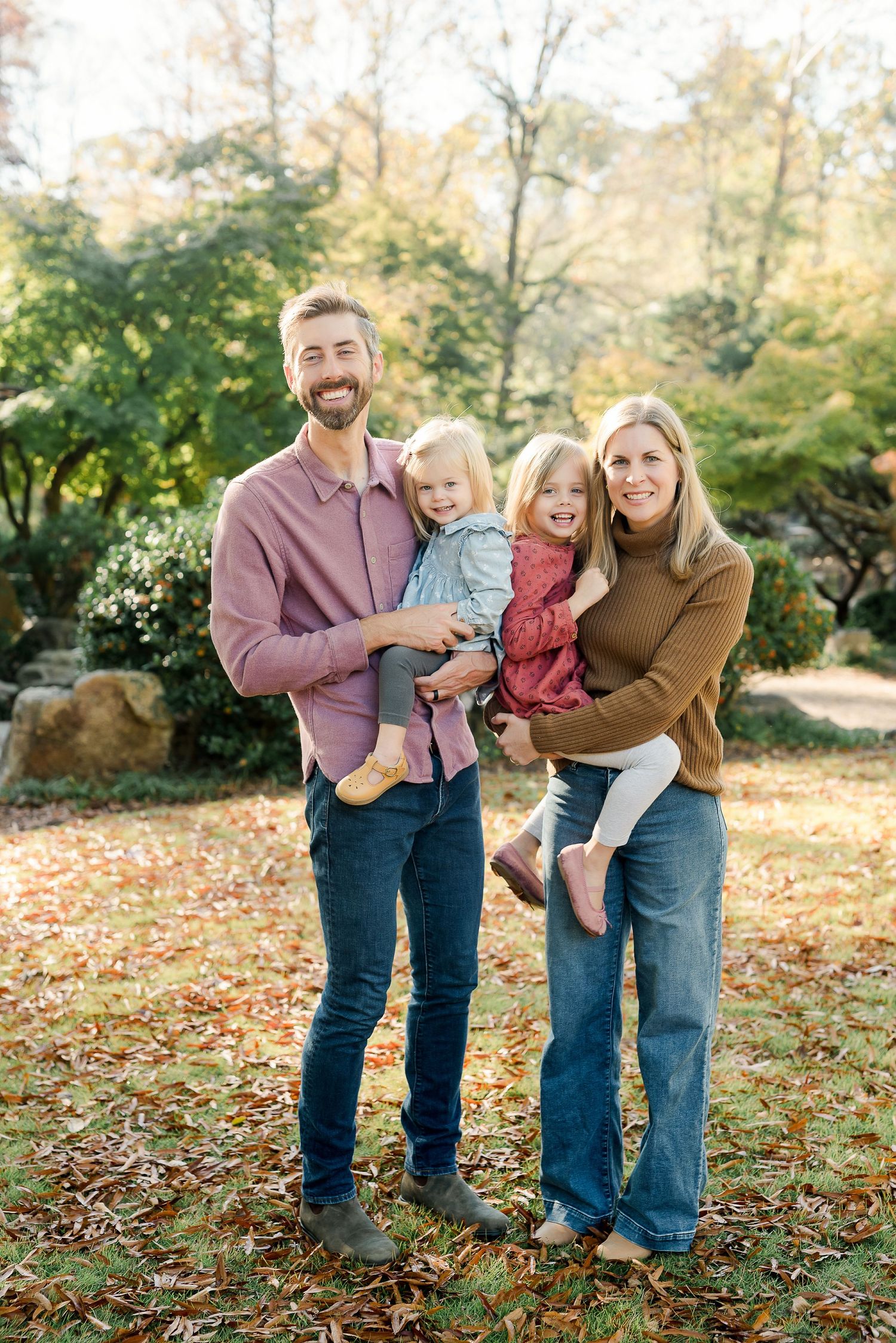 A smiling family of four poses outdoors in fall foliage, parents holding two young children in a sunlit park.