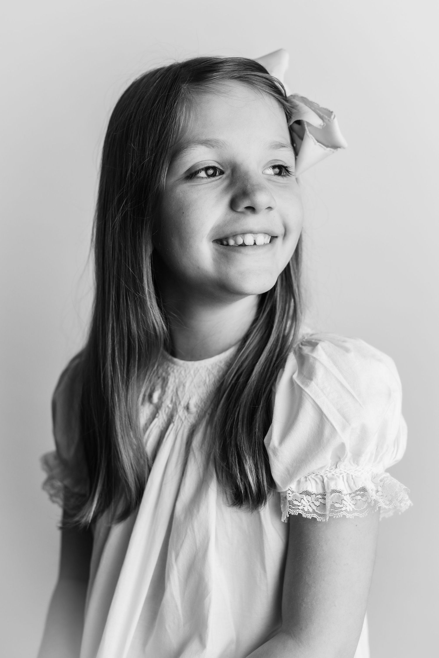 Black and white studio portrait of young person in white dress with bow accessory.