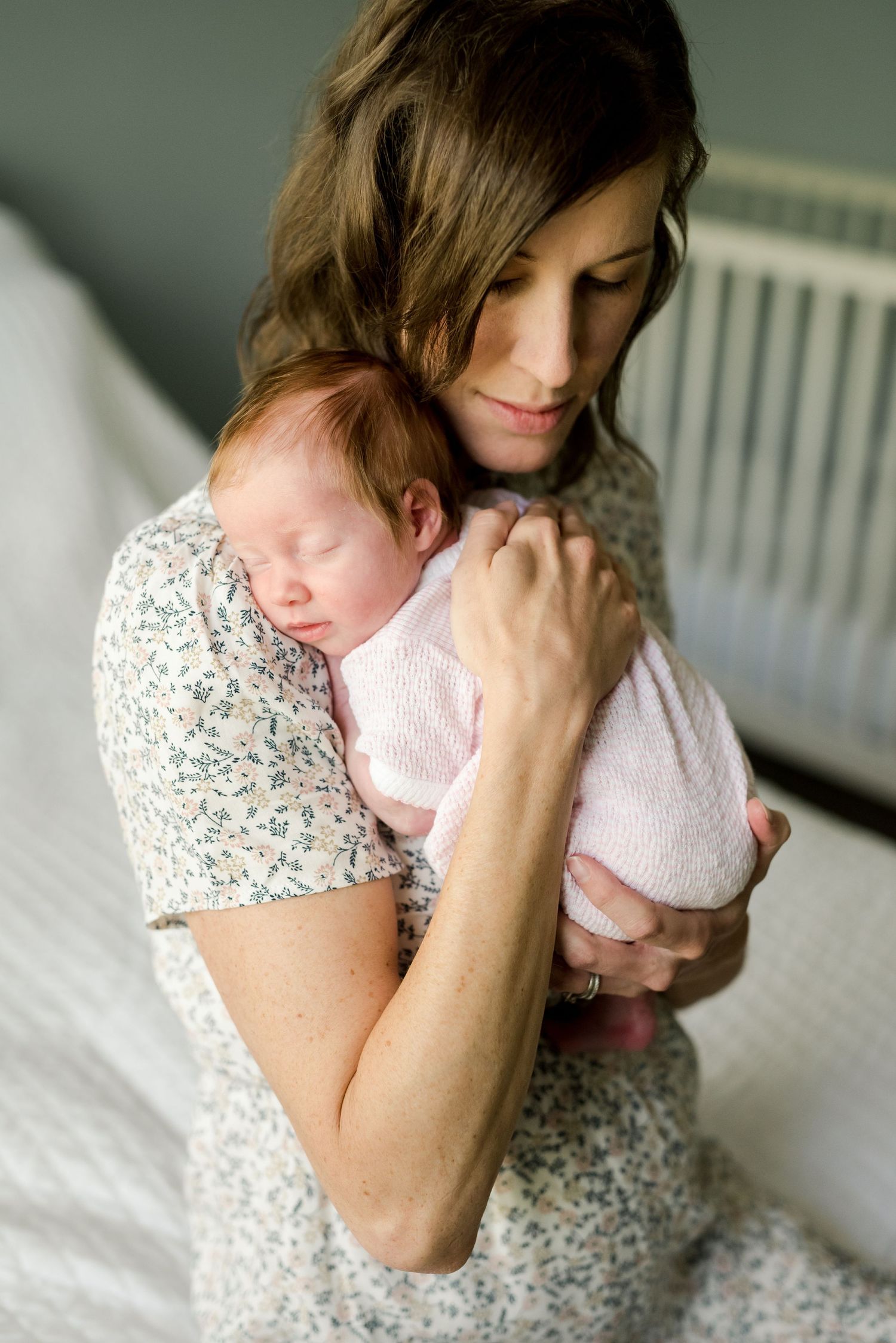 Intimate indoor portrait of mother cradling sleeping infant against gray nursery background.