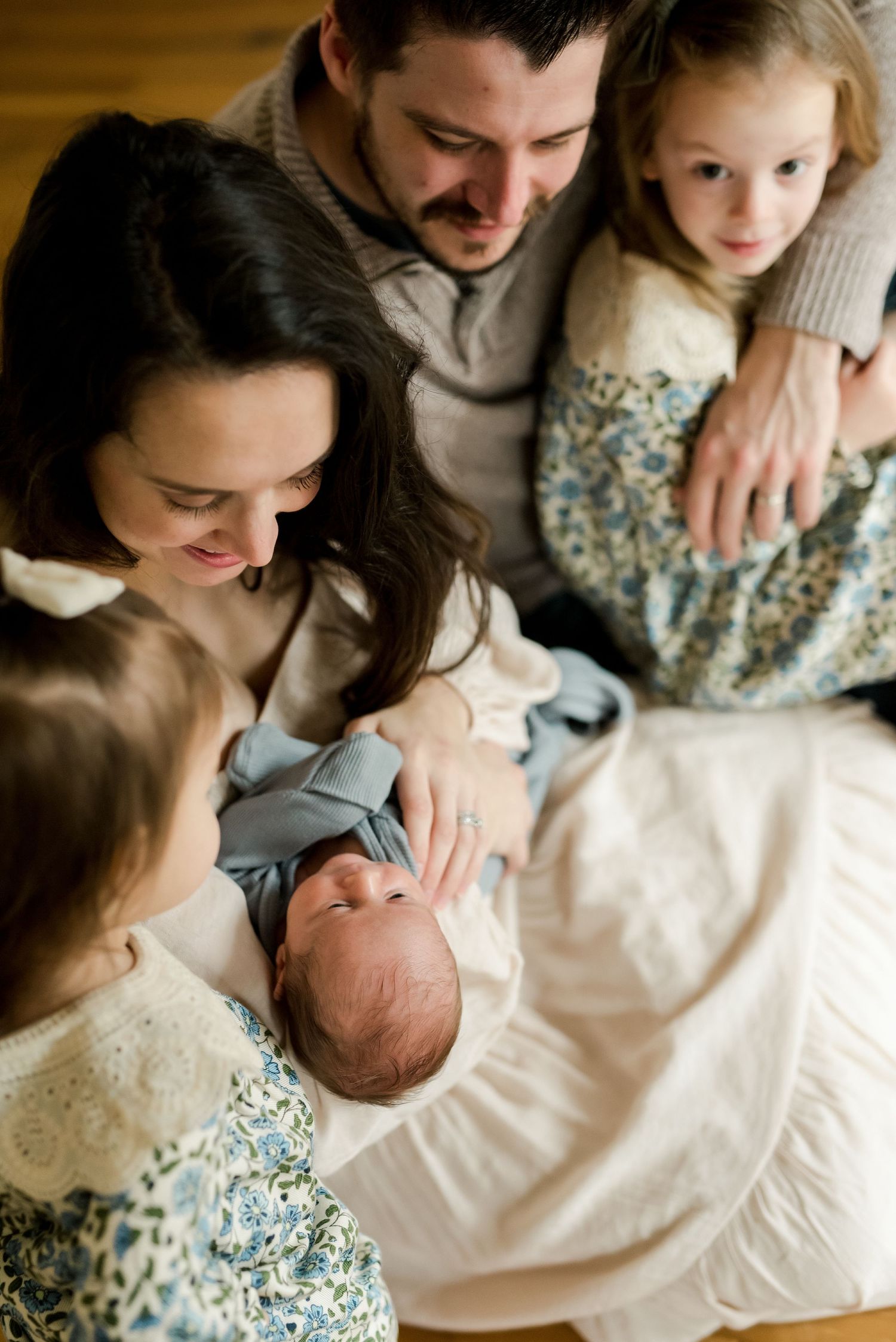 Family sharing intimate moment with newborn baby on hardwood floor.