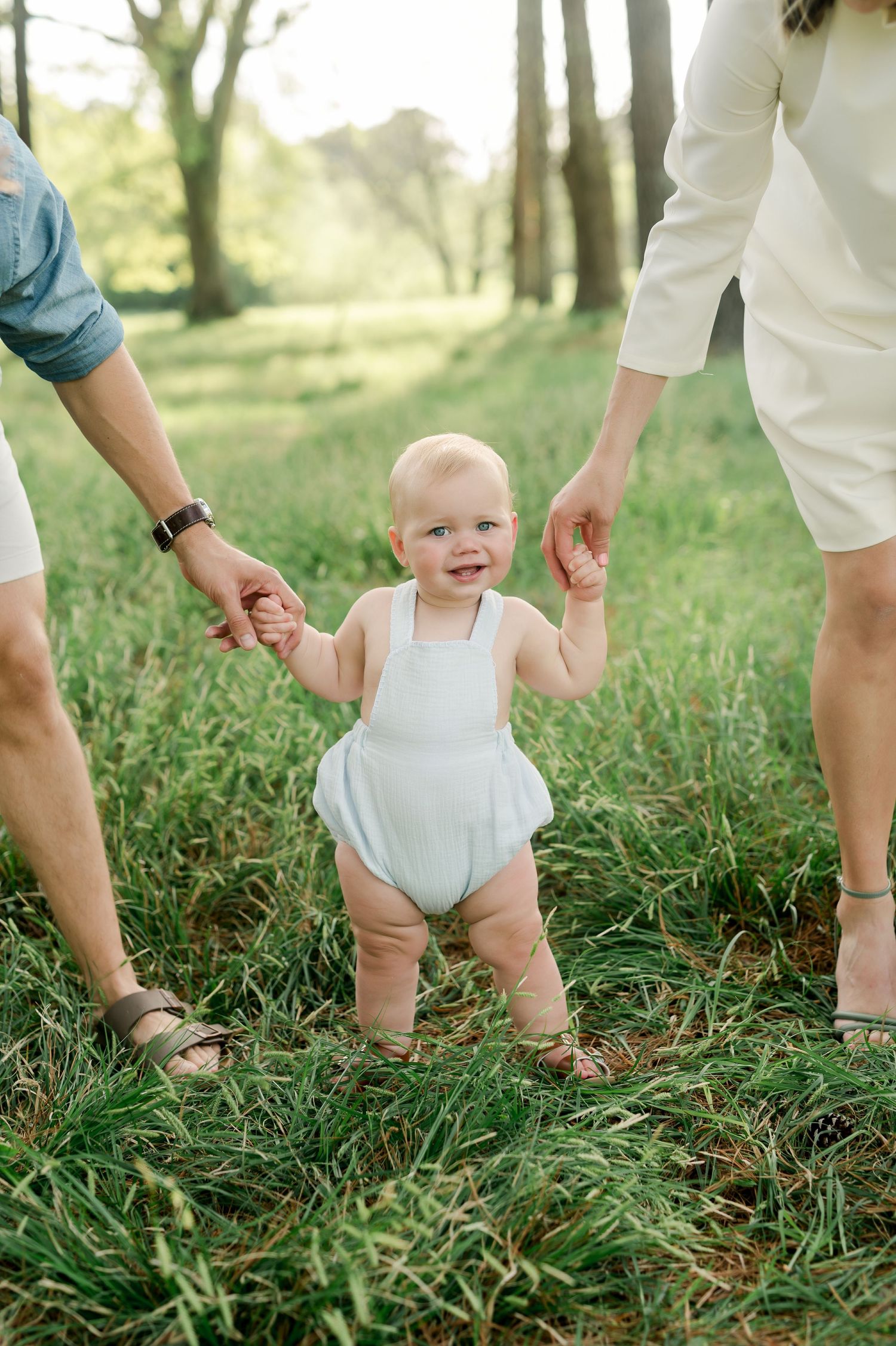 A smiling baby in a white romper stands in green grass while holding hands with adults on a sunny day.