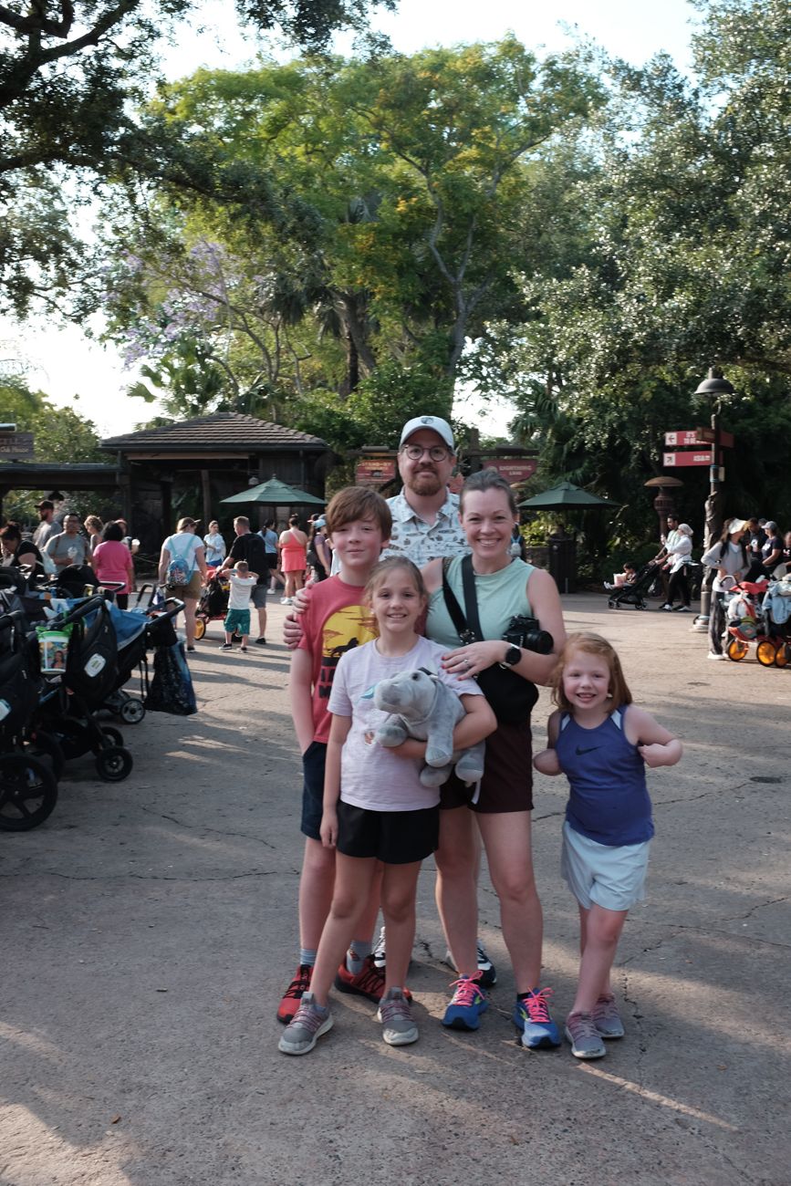 Family group poses together on pathway at Disney theme park with strollers visible in background.