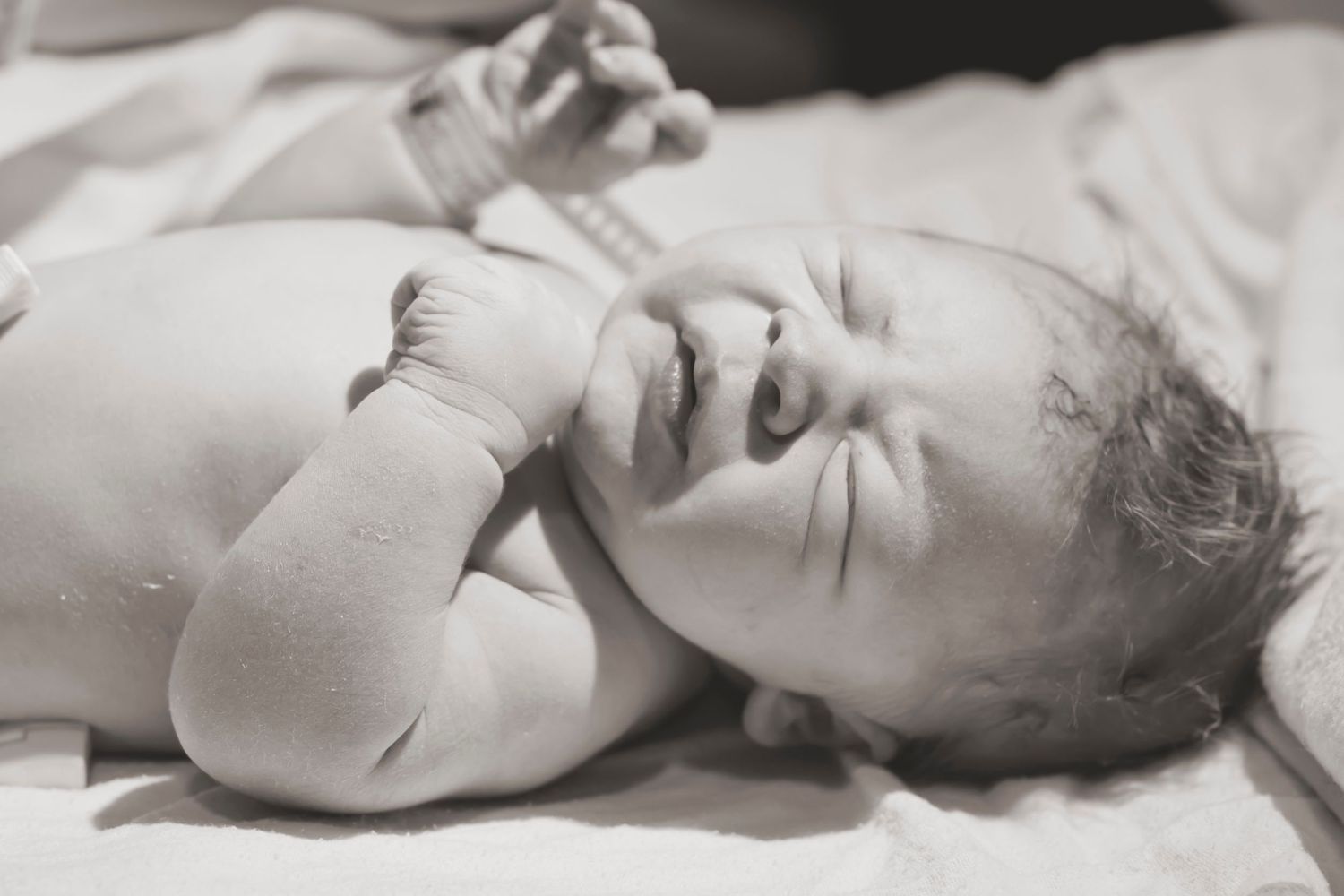 Black and white portrait of an infant lying on soft bedding with tiny hands raised.