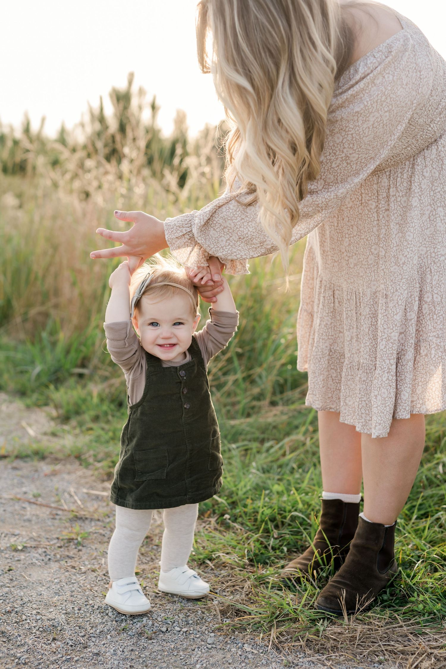 Mother helping toddler walk on dirt path with grass and sunlight in background.