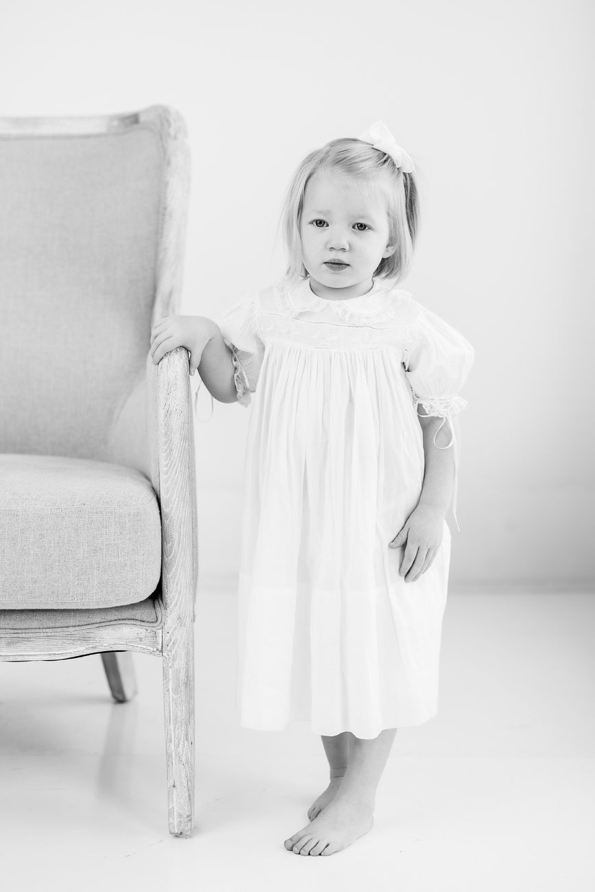 Black and white photo of child in white dress standing barefoot next to gray chair.